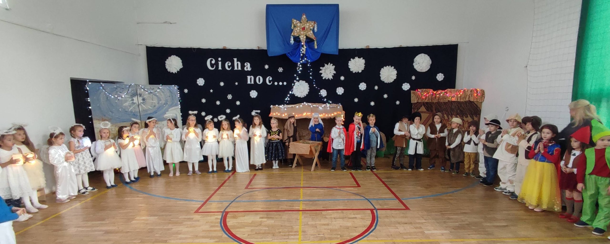 W sali gimnastycznej na tle dekoracji świątecznej z napisem „Cicha noc...”, stoją uczniowie klas 0A i 0B. Ubrani w stroje pastuszka, aniołka, postaci z bajek.