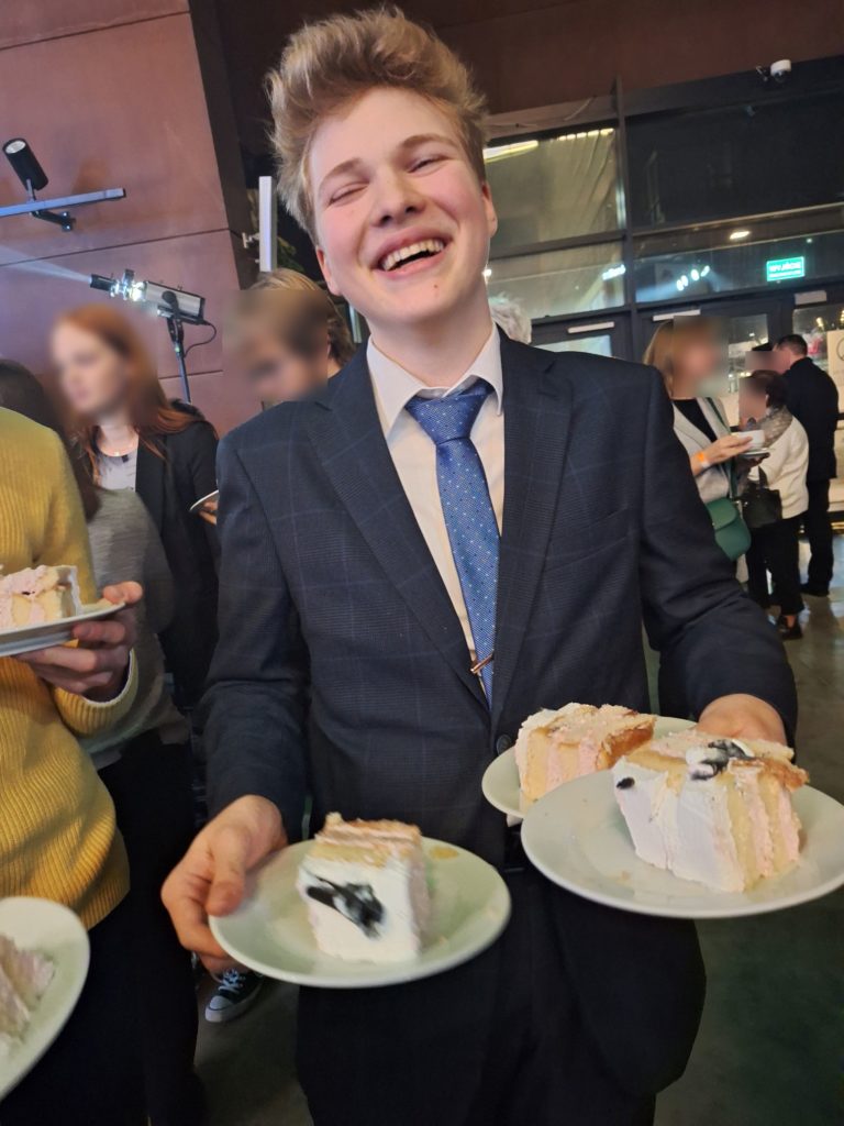 Chłopak z wielkim uśmiechem ubrany w garnitur stoi i trzyma w rękach 3 talerzyki z ciastem.