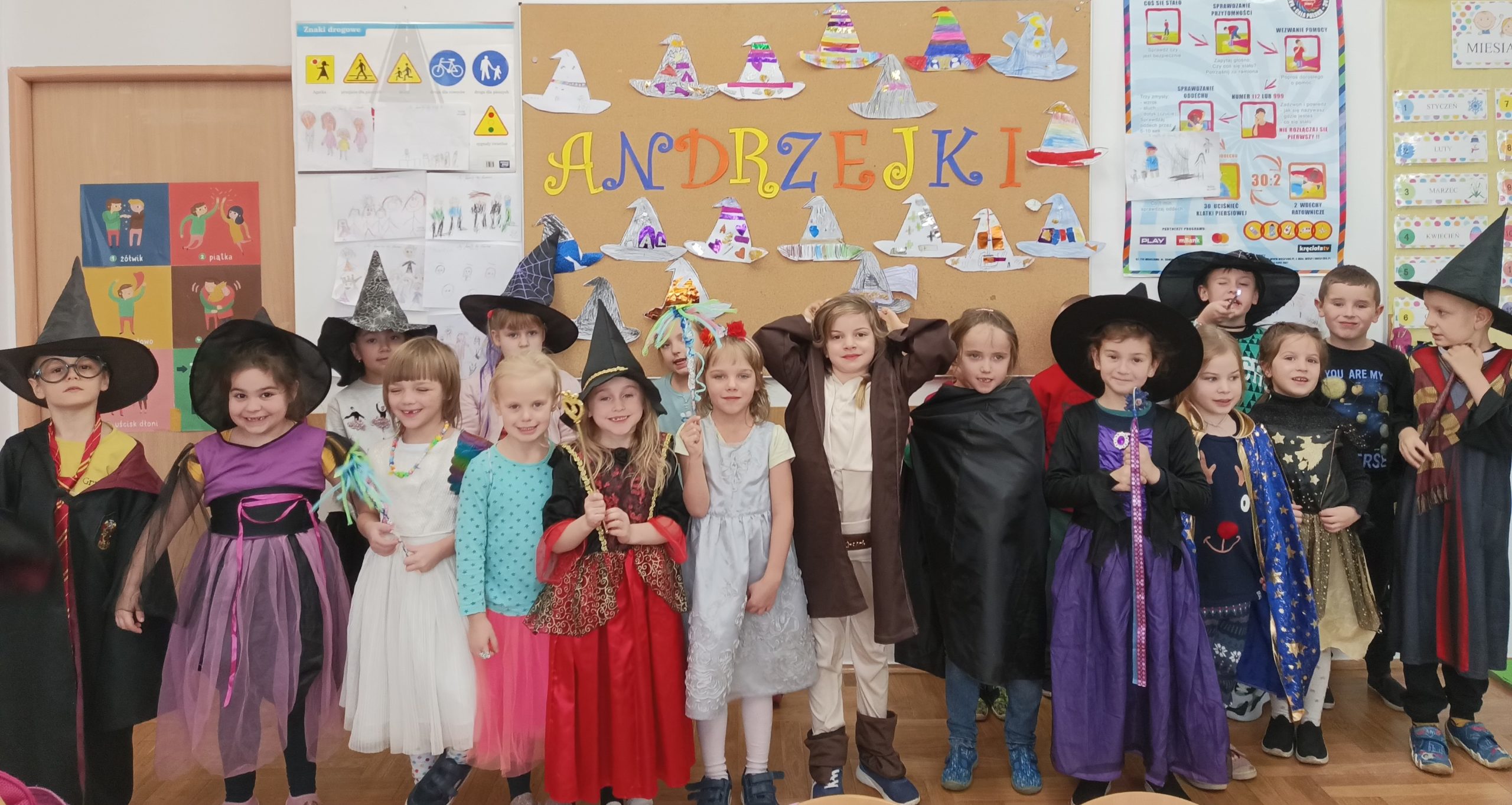  Dzieci stoją z uśmiechem na twarzy, ubrani w stroje wróżek, magików, czarodziei. W tle przyczepione do tablicy korkowej girlandy z napisem Andrzejki oraz kolorowe czarodziejskie czapki.