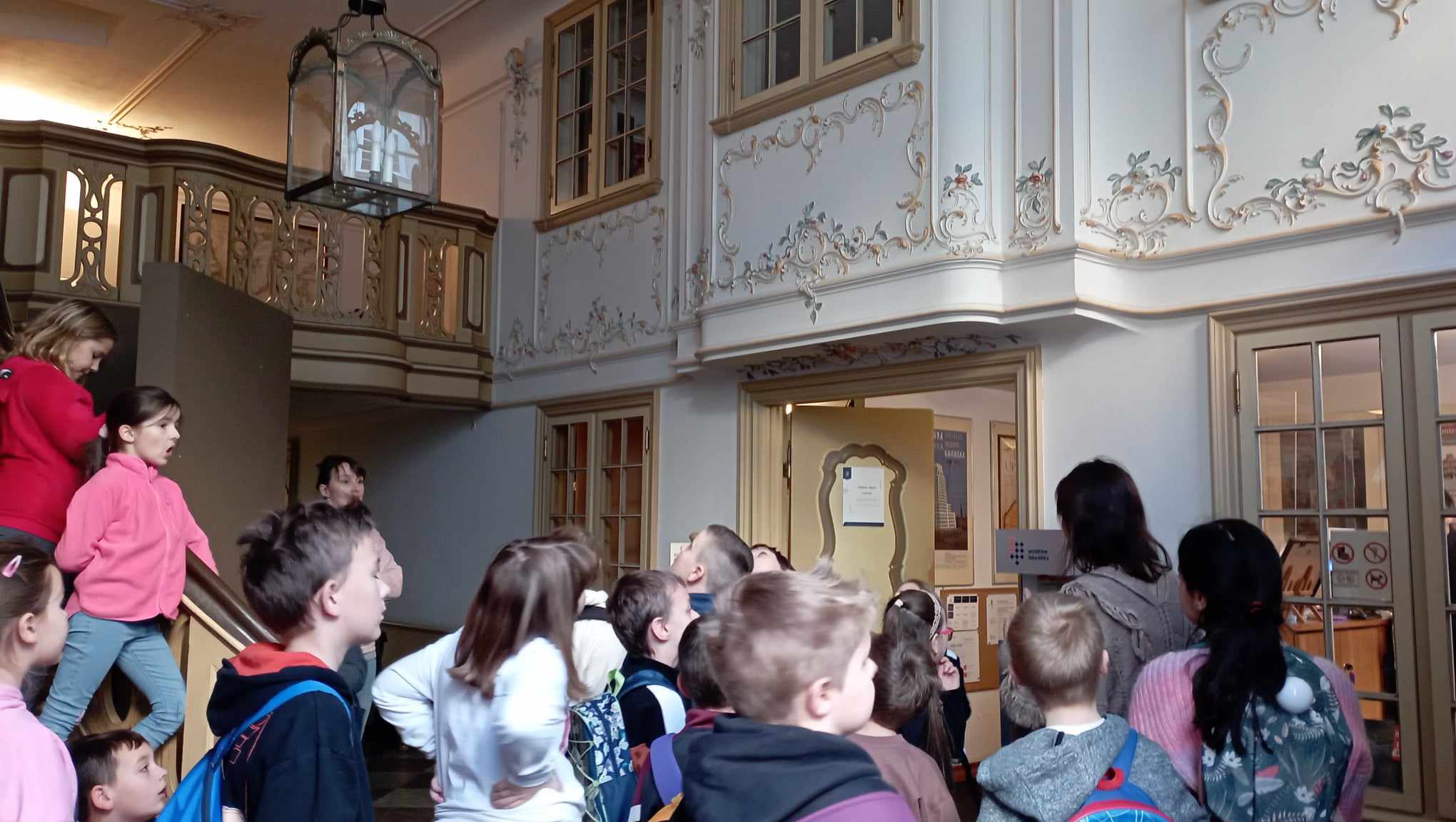 W sieni Domu Uphagena stoją uczniowie klasy 2c wraz z przewodniczką. W tle schody, antresola i ozdobna lampa.
