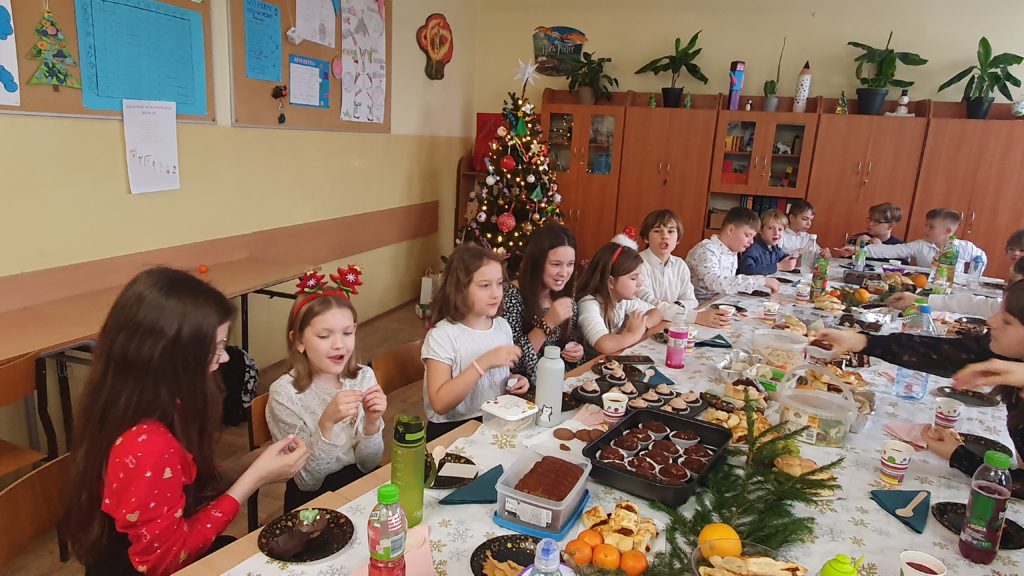 Grupa kilkunastu uczniów siedzących przy wigilijnym stole. Na stole jest wiele potraw: ciasta, owoce, sałatki. w tle choinka ubrana w kolorowe ozdoby.