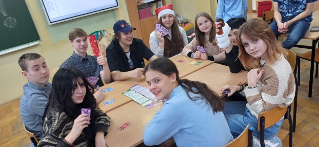 6 dziewczyn i 3 chłopców siedzą przy stole, trzymając karty do gry Wirus. Są uśmiechnięci i zadowoleni.