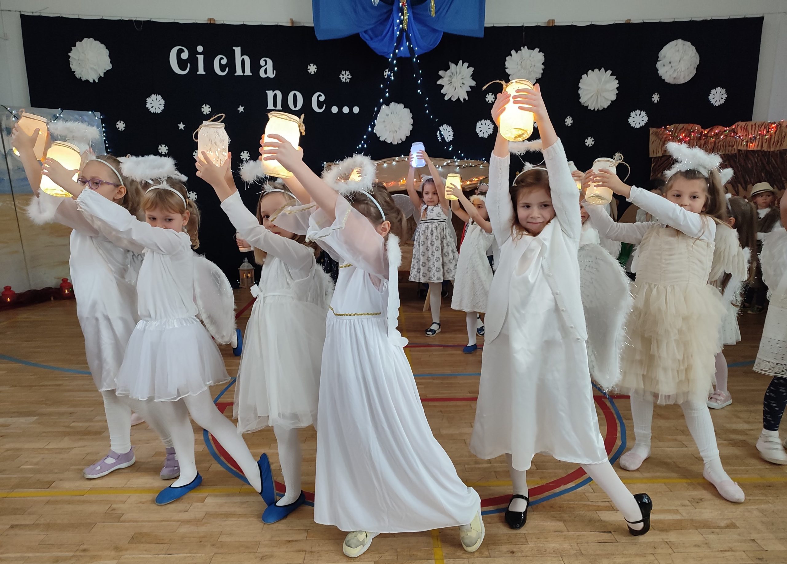 W sali gimnastycznej na tle dekoracji świątecznej z napisem „Cicha noc...”, na pierwszym planie tańczą ucze nnice ubrane w strój aniołka. Trzymają w rękach lampiony.