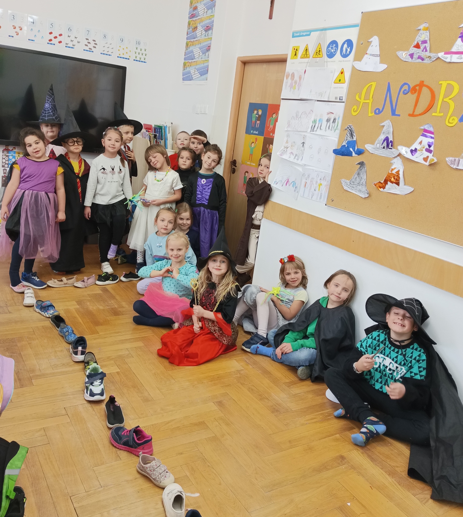  Dzieci stoją/siedzą uśmiechem na twarzy, ubrani w stroje wróżek, magików, czarodziei. Na podłodze ułożone buty w rzędzie.