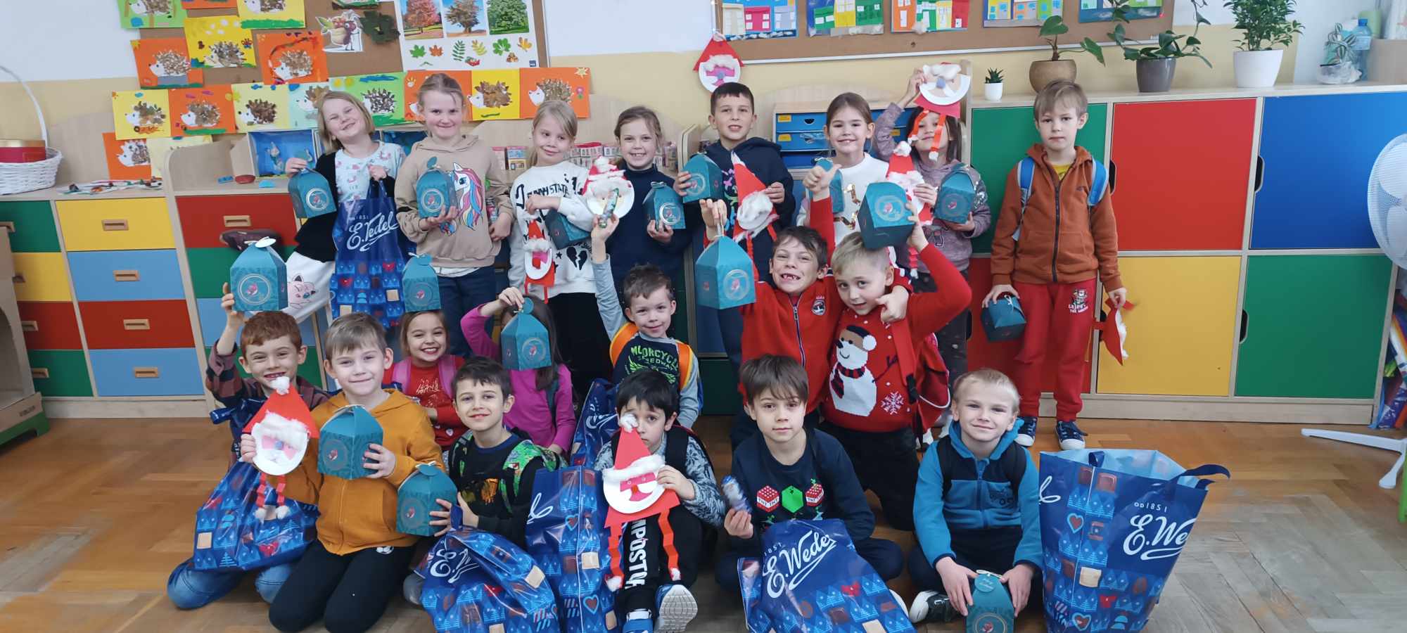 Sala szkolna, uczniowie ustawieni w trzech rzędach trzymający papierowe Mikołaje i niebieskie torby z napisem Wedel.