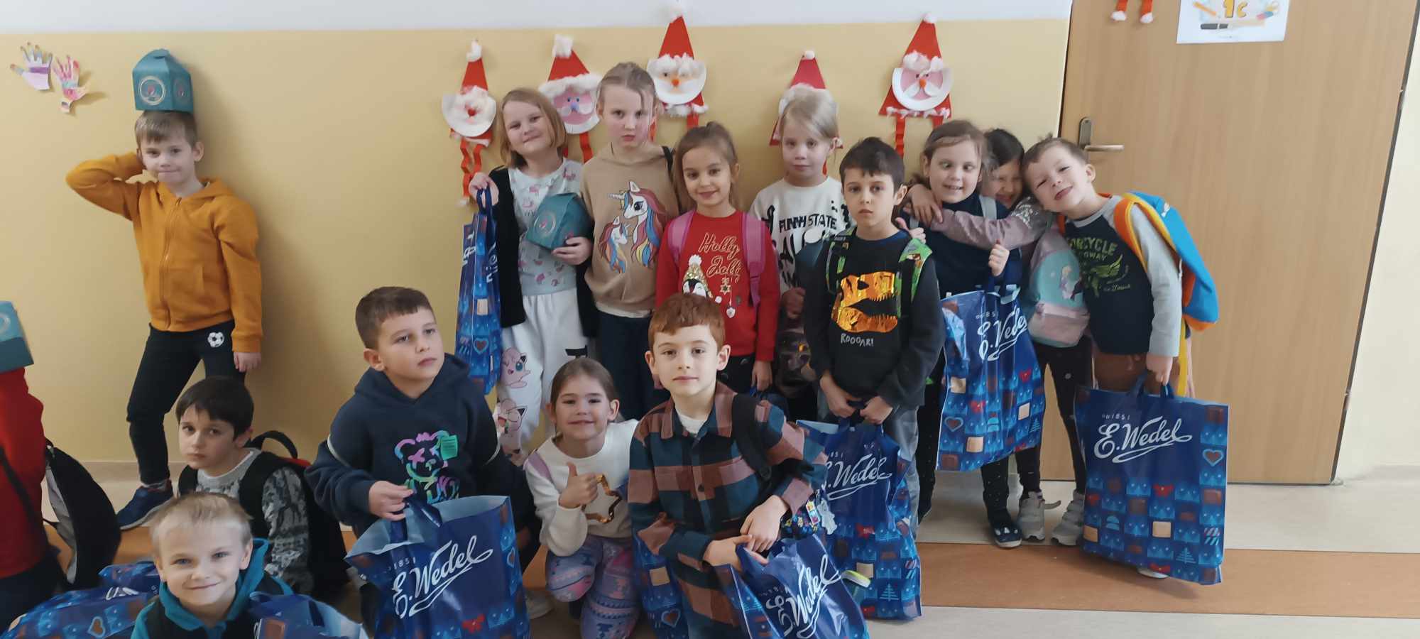 Sala szkolna, uczniowie ustawieni w trzech rzędach trzymający papierowe Mikołaje i niebieskie torby z napisem Wedel.