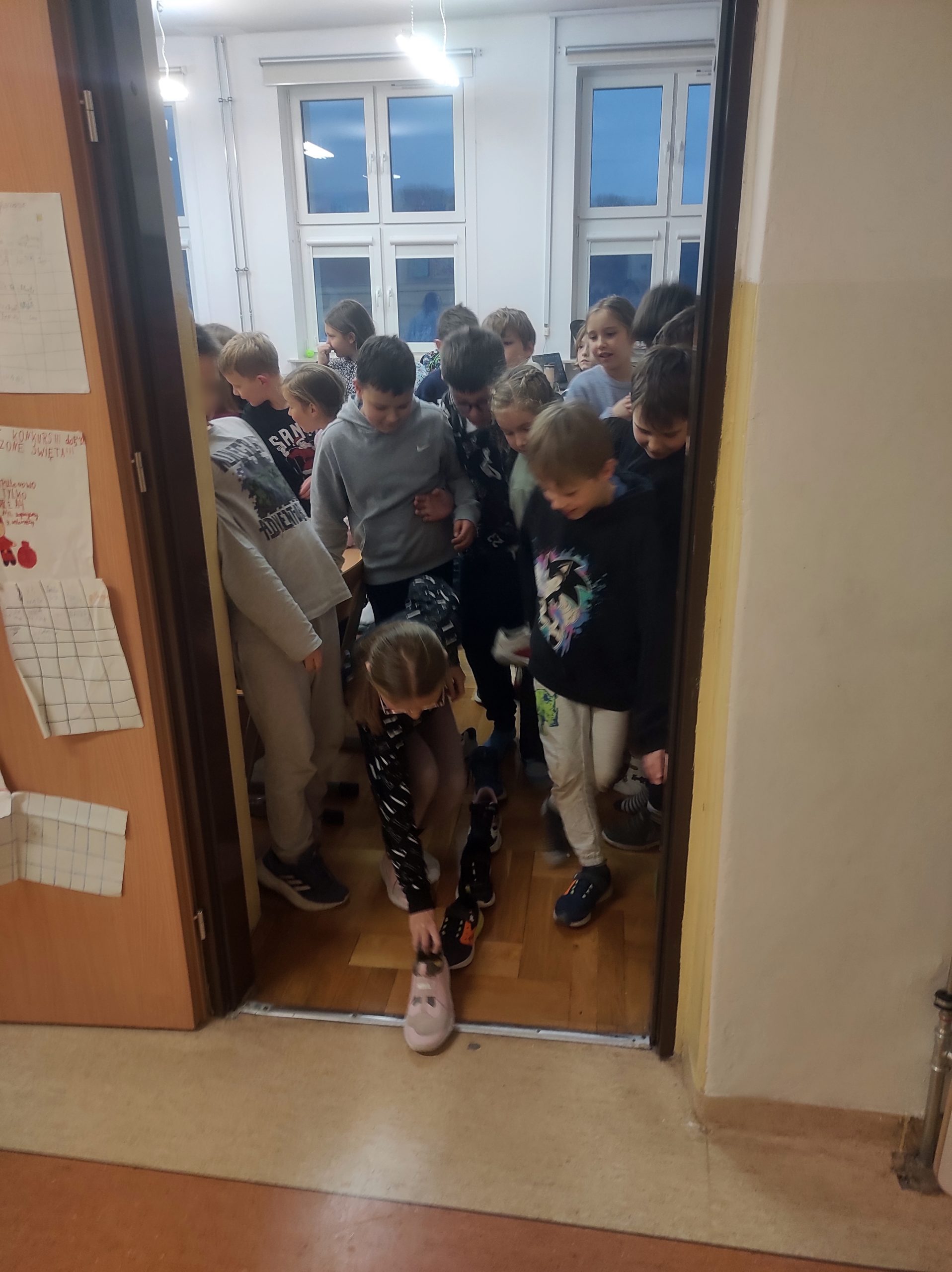 W drzwiach stoi grupa uczniów 3b, na pierwszym planie nad szpalerem z butów pochyla się dziewczynka, która kładzie swojego buta na progu.