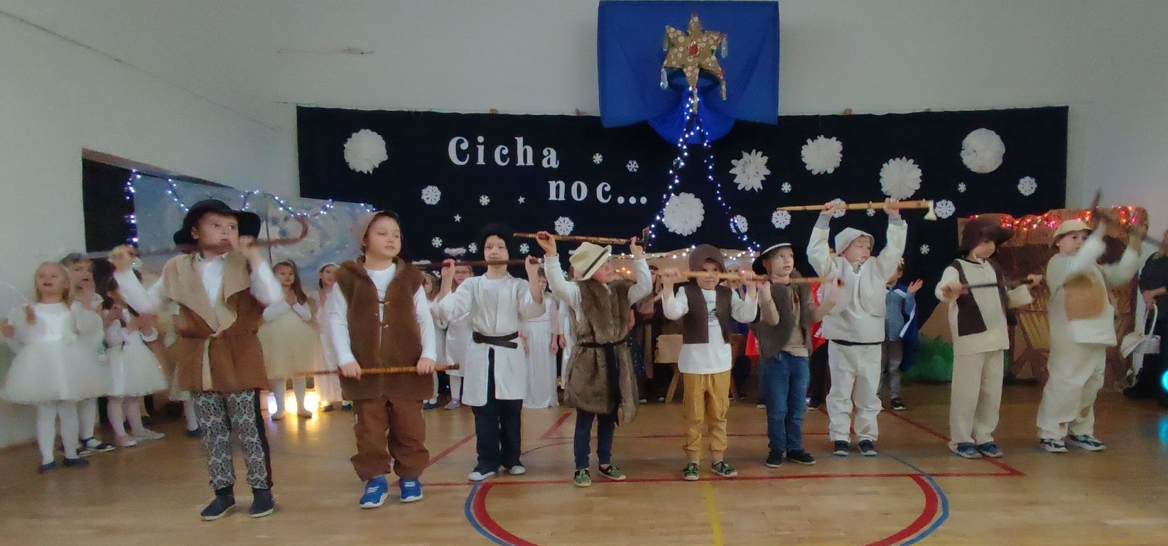 W sali gimnastycznej na tle dekoracji świątecznej z napisem „Cicha noc...”, na pierwszym planie stoją uczniowie ubrani w strój pastuszka. W rękach trzymają ciupagi.