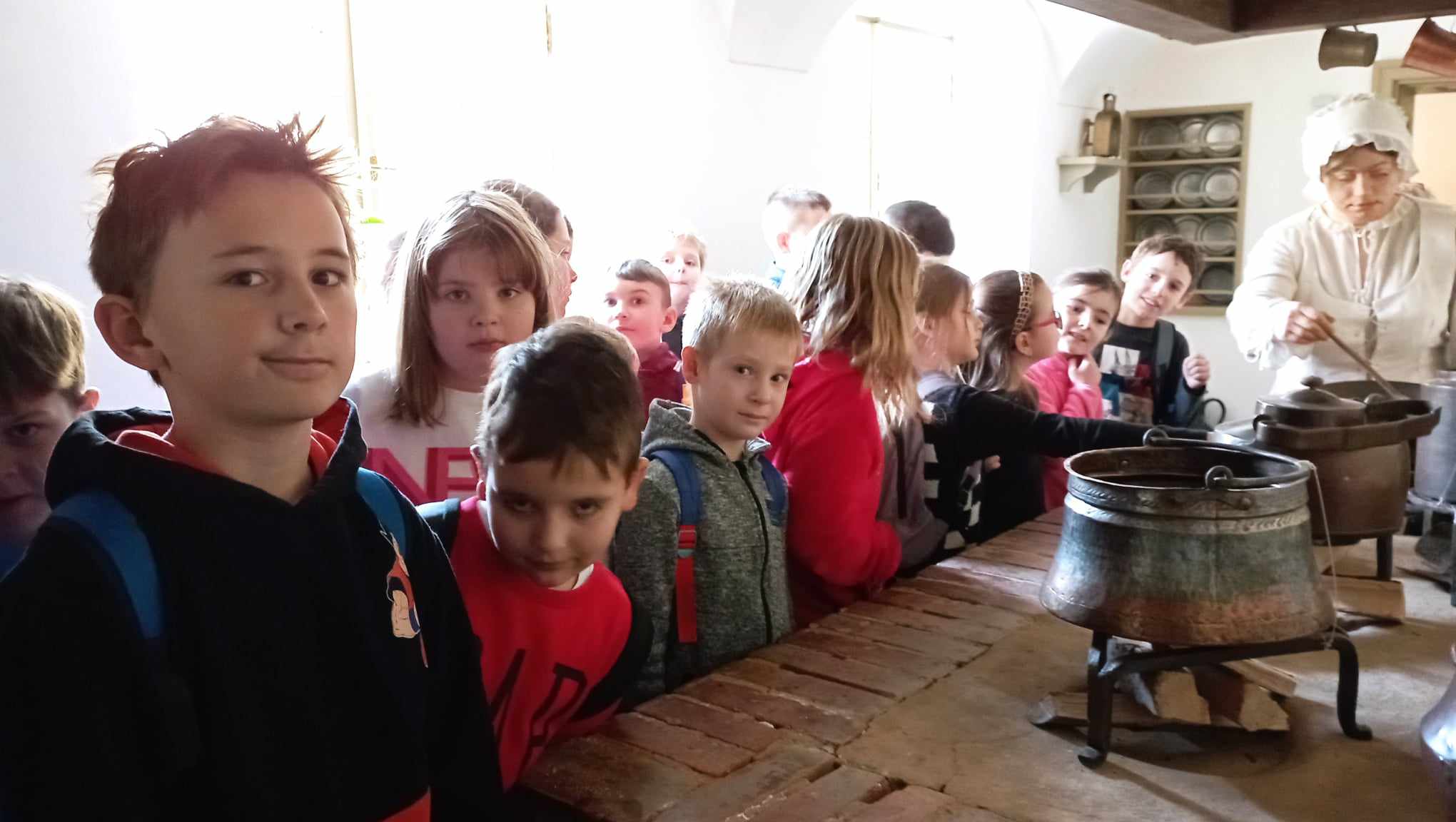 W zabytkowej kuchni Domu Uphagena stoją uczniowie klasy 2c. W tle figura kucharki, regał z naczyniami oraz dwa kociołki.