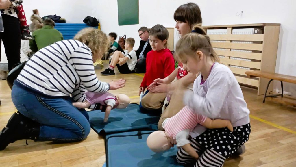 W sali gimnastycznej na pierwszym planie uczennica i kobieta uderzające fantomy niemowląt między łopatki, obok dwie inne uczennice. W tle inni rodzice i uczniowie siedzący na podłodze.