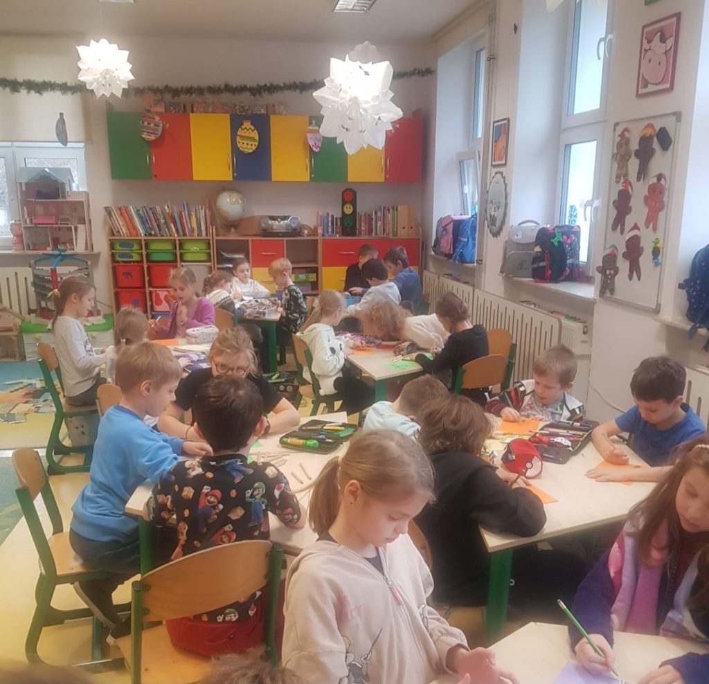  Ok 25 uczniów siedzi przy stolikach w sali świetlicy szkolnej, przed sobą mają kolorowe kartki oraz piórniki z kredkami i mazakami.