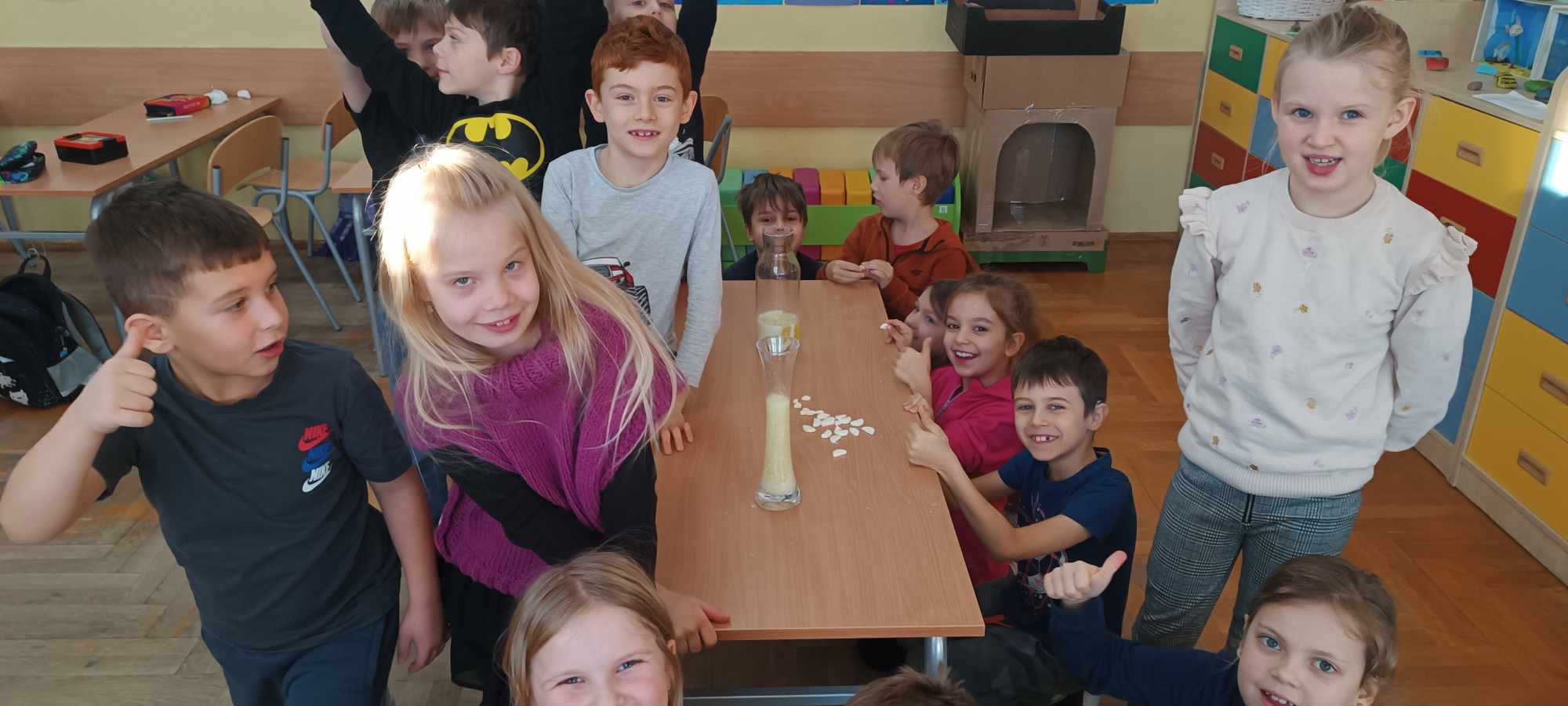 Grupa dzieci w klasie szkolnej stojąca wokół stołu. Na stole dwa szklane naczynia z kolorową cieczą.