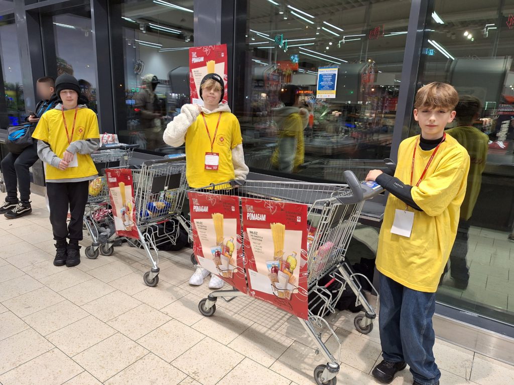 3 chłopców w koszulkach wolontariuszy stoi w sklepie przy dwóch wózkach sklepowych, na których wiszą plakaty dotyczące zbiórki żywności. Na szyjach mają zawieszone identyfikatory.