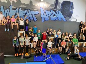 grupa uczniów w strojach sportowych siedzi lub stoi na tle ścianki z napisem Movement Arena