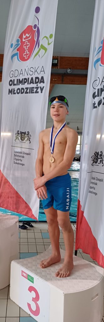 Chłopiec stoi na podium z nr 3, w tle znajdują się flagi z napisem „Gdańska Olimpiada Młodzieży”. Ubrany jest w strój pływacki – niebieskie spodenki do kolan, czepek i na nim czarne okularki pływackie. Na szyi zawieszony ma medal na biało niebieskiej wstążce koloru brązu