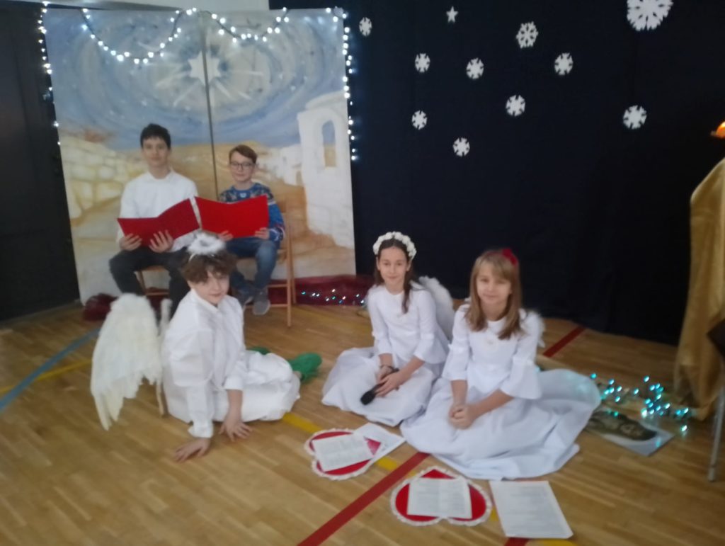 3 dziewczynki w strojach aniołów siedzą na podłodze i 2 chłopców siedzi na krześle trzymając w ręku teczki. Jedna dziewczynka trzyma w ręku mikrofon.