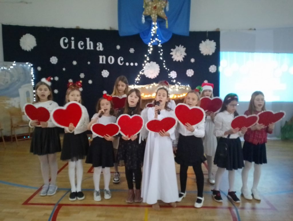 11 dziewczynek ubranych galowo stoją na środku sali i trzymają w ręku duże papierowe serca. Jedna z nich ubrana jest w stój anioła i śpiewa do mikrofonu.