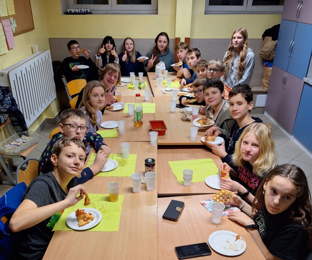 Grupa dzieci siedzi wokół długiego stołu i jedzą pizze. Na ich twarzach jest uśmiech i zadowolenie.