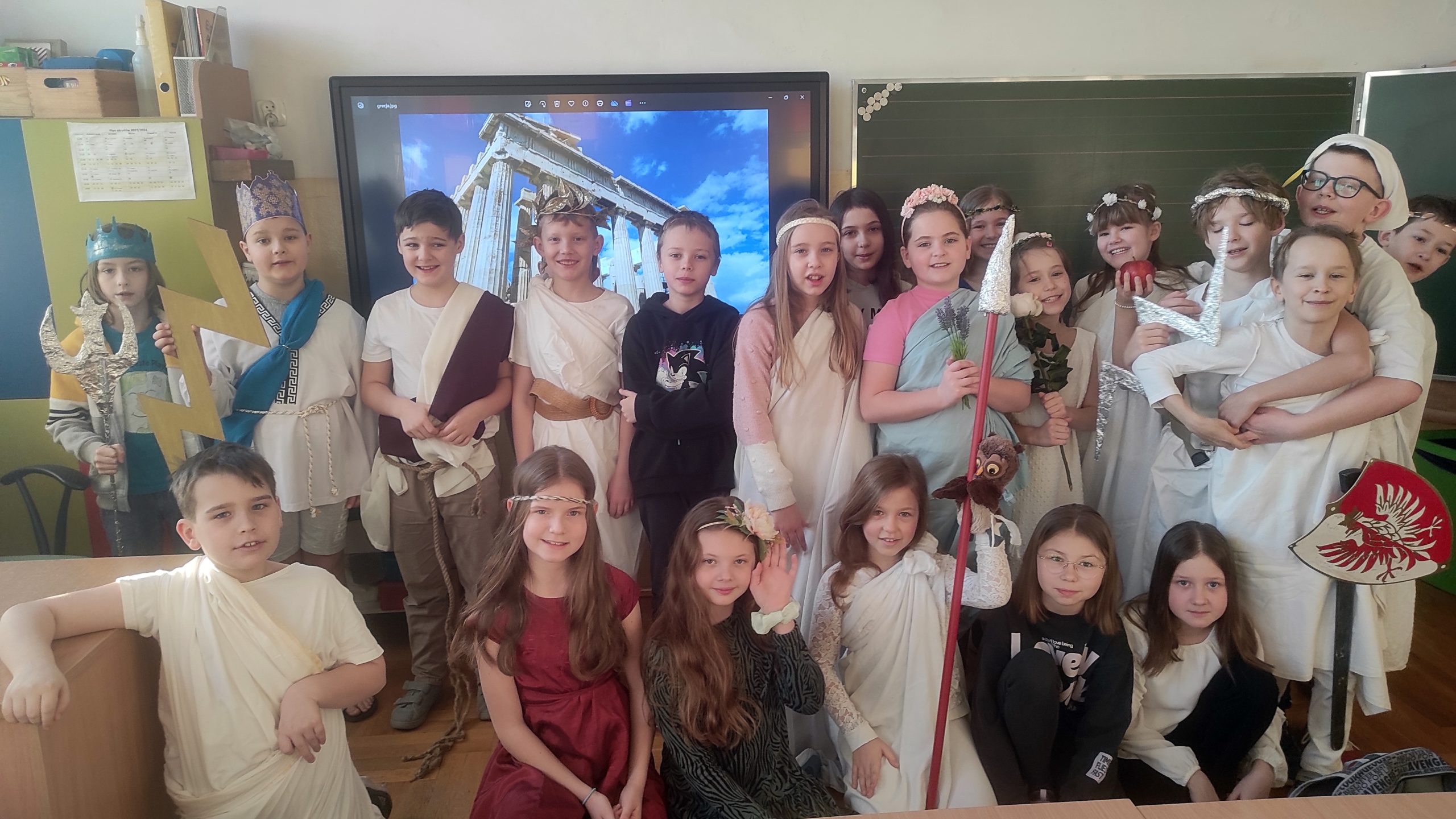 Grupa 21 uczniów przebranych za boginie i bogów z Olimpu ustawiona w 2 rzędach. Pierwszy rząd z tyłu stoi, przed nimi dzieci klęczą. W tle Partenon wyświetlony na telewizorze.