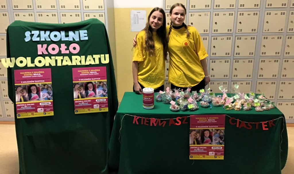 Dwie dziewczynki w żółtych bluzkach stoją przy stole, na stole leżą zapakowane w folię babeczki, po lewej stronie stoi tablica, na której jest napis Szkolne koło wolontariatu i dwa plakaty o akcji charytatywnej
