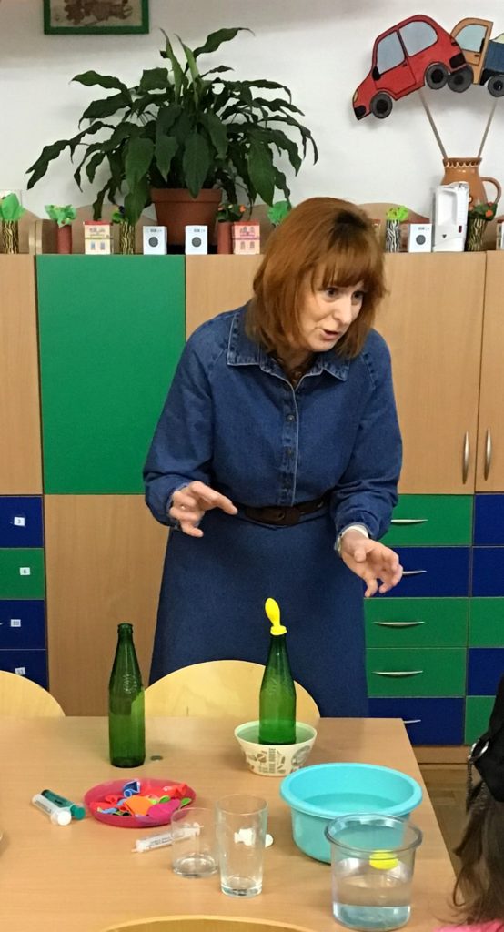 Nauczycielka w niebieskiej sukience stoi przy stole i prezentuje doświadczenie z butelkami szklanymi i balonem. Na stole przed nią leżą niebieska miska, czerwony talerzyk z balonami oraz szklane naczynia z wodą. W tle stoi szafa z niebieskimi i zielonymi frontami, na szafie stoi kwiat