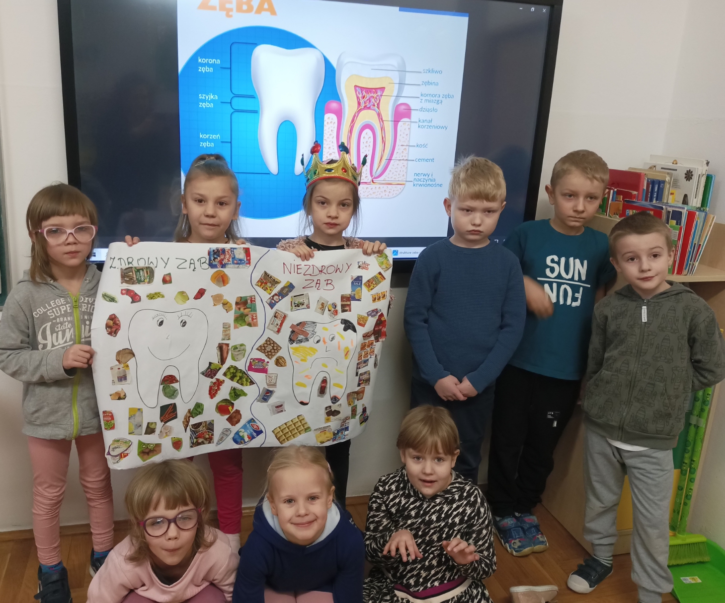9 uczniów znajduje się w sali lekcyjnej. Dzieci trzymają w rękach plakat, który przedstawia produkty spożywcze, które mogą niszczyć zęby oraz produkty, które pomagają mieć zdrowe zęby.