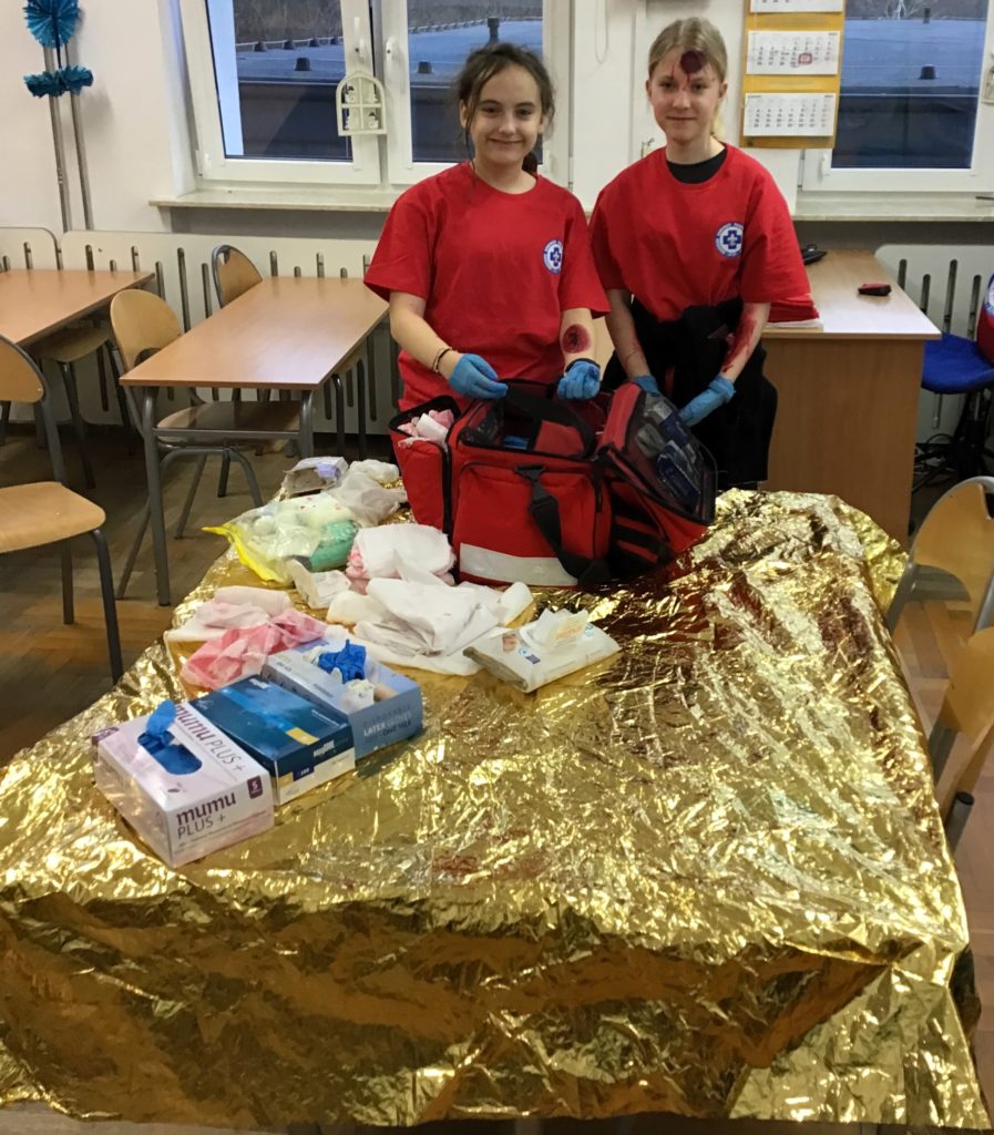 Dwie dziewczynki ubrane w czerwone koszulki stoją przy stole, na którym leżą przedmioty służące do udzielania pierwszej pomocy przedmedycznej. Dziewczynki są pomalowane na twarzy, symulując ranę