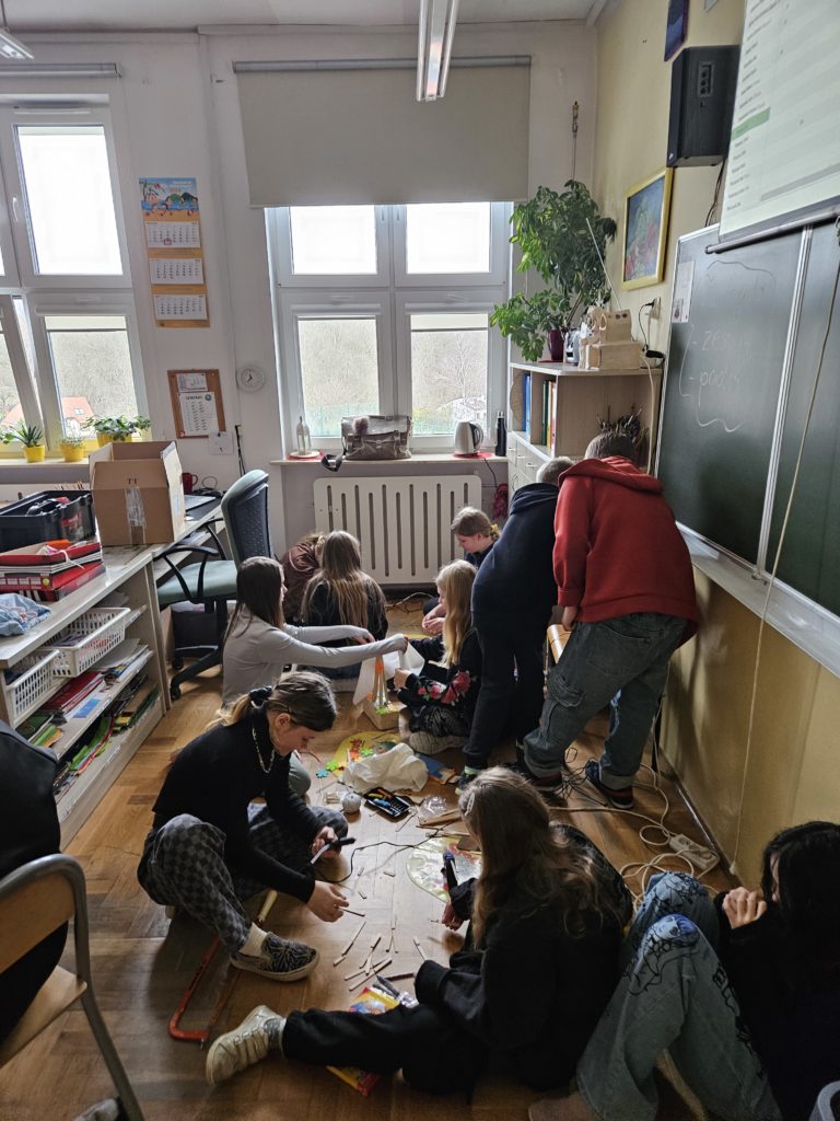 Uczniowie na podlodze skupieni przy swoich pracach z drewna. Sklejają i montują drewniane elementy