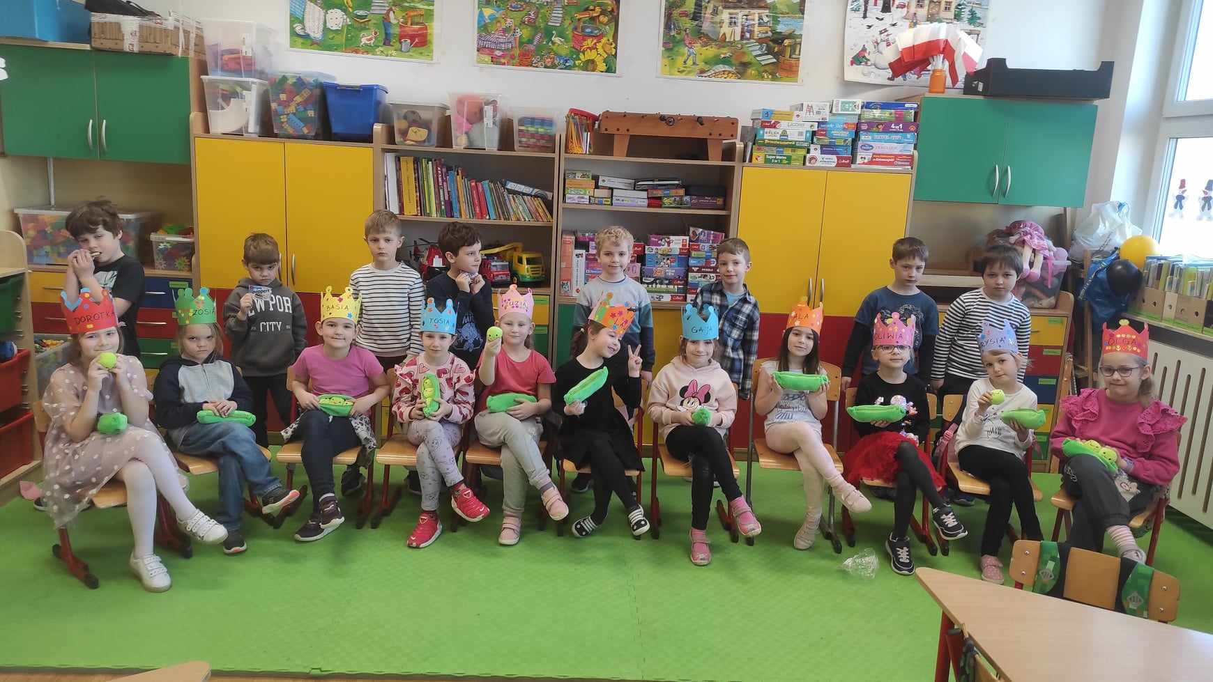 8 chłopców stoi, a 11 dziewczynek siedzi na krzesłach w sali lekcyjnej. Dziewczynki trzymają w rękach zielone piórniki, a na głowie mają papierowe korony. W tle szafki i zabawki.