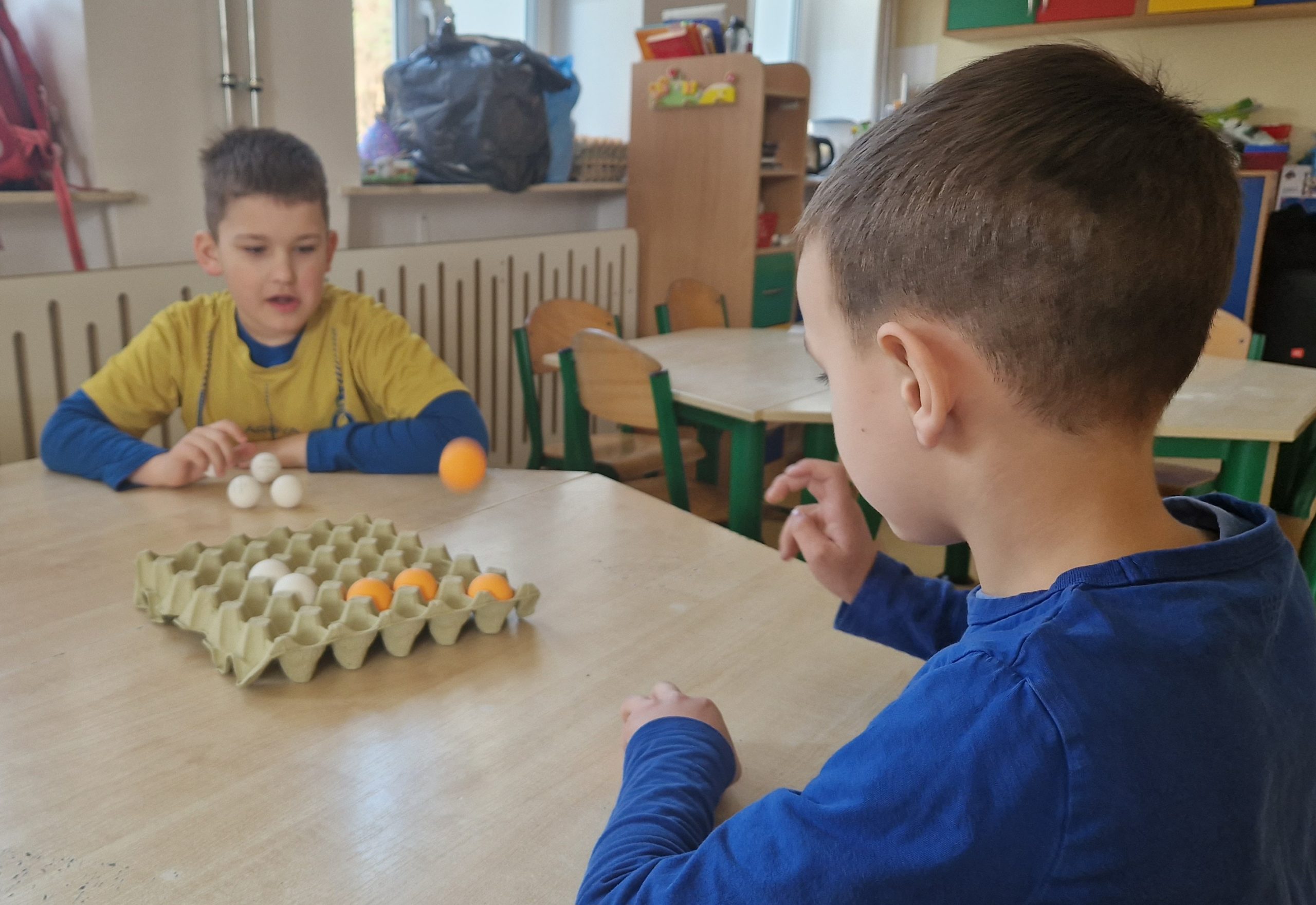 Przy stoliku siedzi 2 chłopców, rzucając piłeczkami do wytłaczanki po jajkach.
