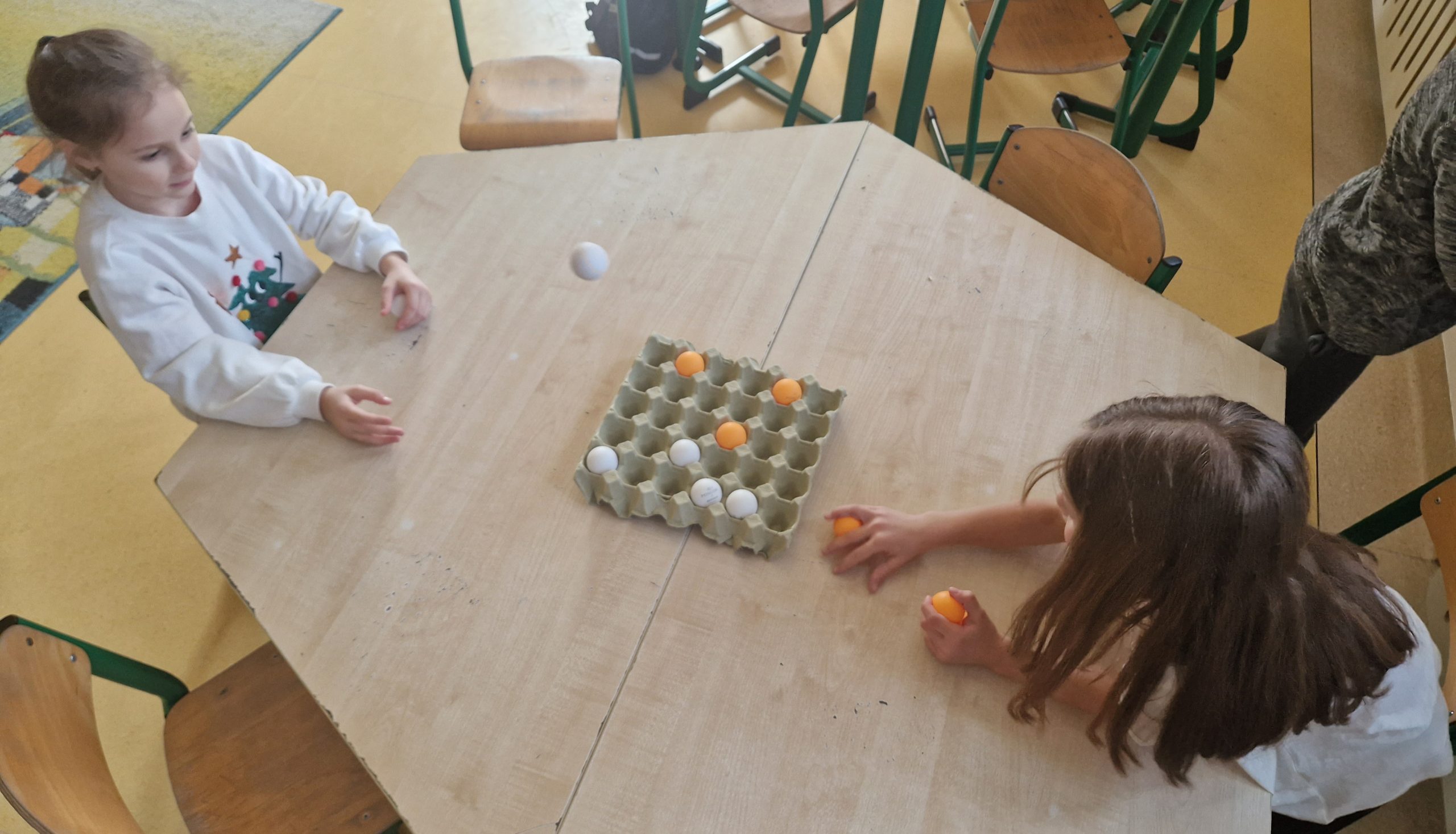 Przy stoliku siedzą 2 dziewczynki, rzucając piłeczki do wytłaczanki po jajkach.