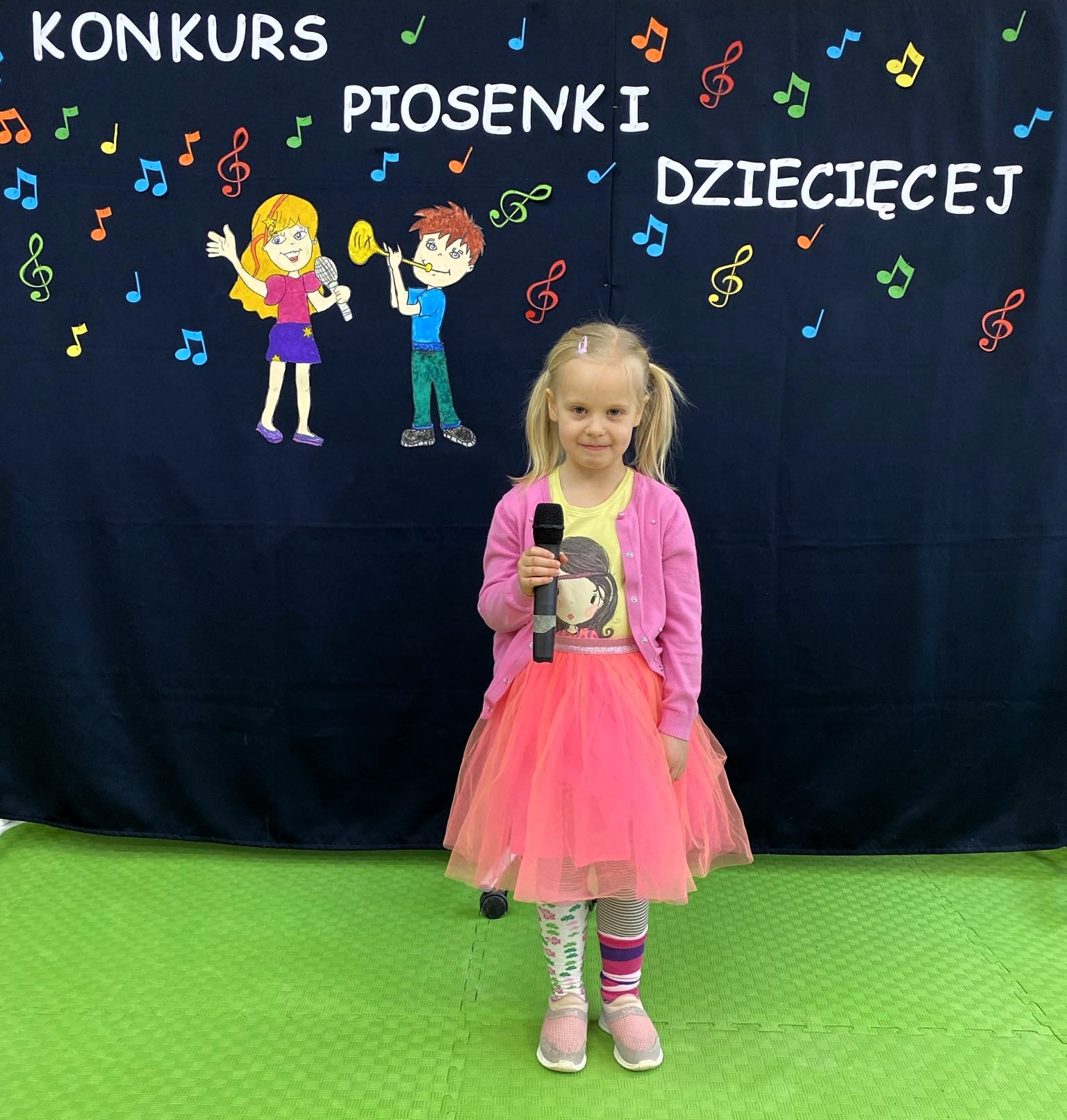 W sali lekcyjnej na tle dekoracji z napisem „Konkurs piosenki dziecięcej”, stoi dziewczynka i trzyma mikrofon.