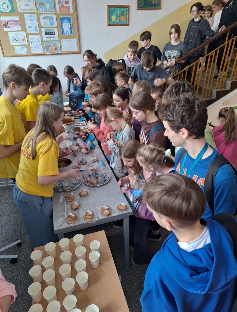 Duża grupa dzieci stoi wzdłuż długiego stołu, na którym lezą półmiski pełne słodkich wypieków. Niektórzy są w trakcie kupowania ciastek.