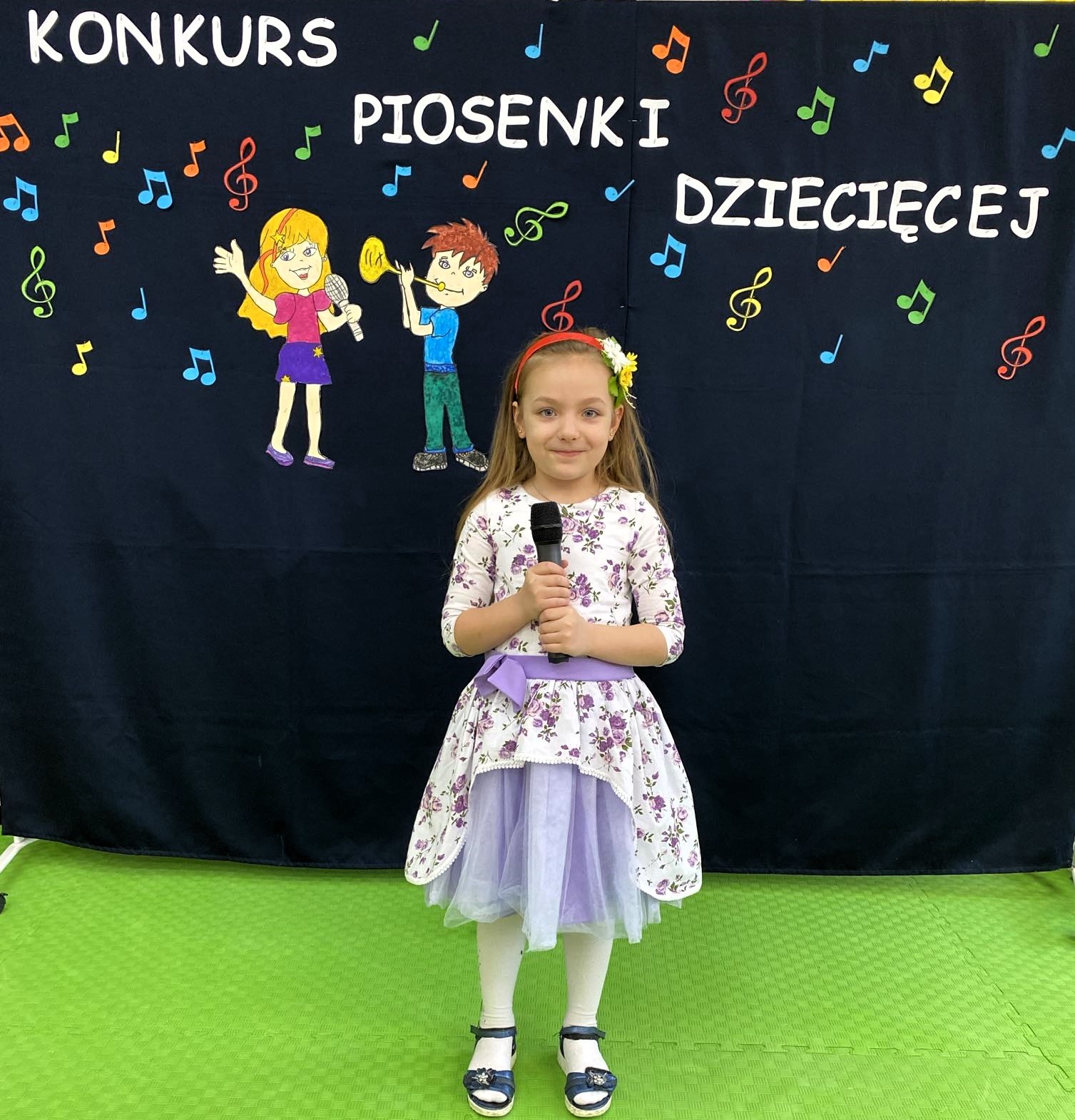 W sali lekcyjnej na tle dekoracji z napisem „Konkurs piosenki dziecięcej”, stoi dziewczynka i trzyma mikrofon.