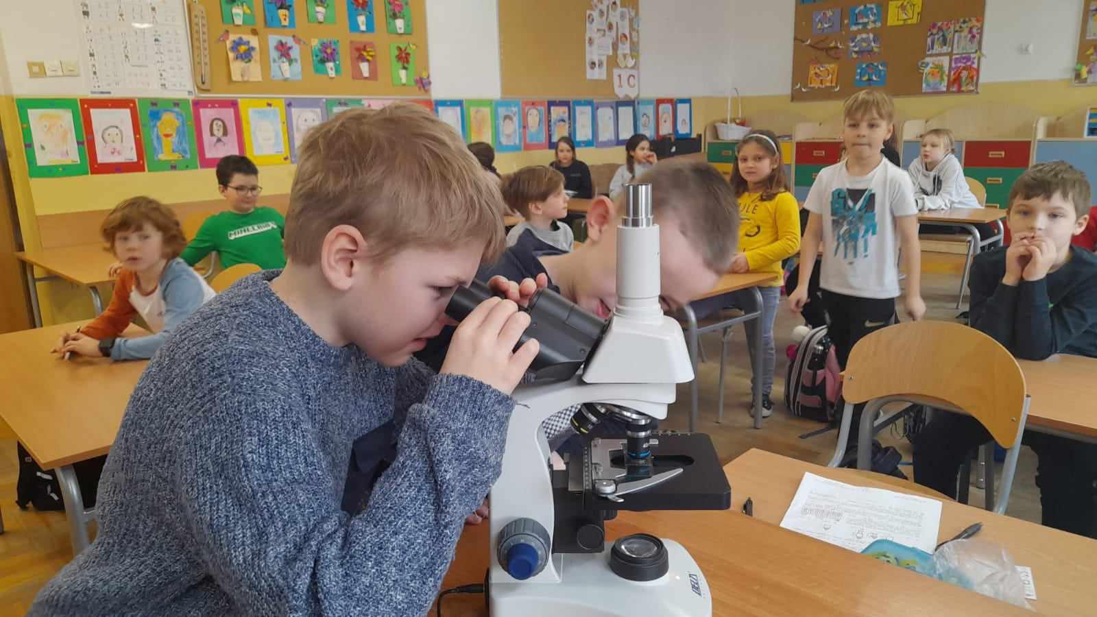 Grupa 20 dzieci znajduje się w sali lekcyjnej. Na pierwszym planie jest chłopiec, który ogląda pod mikroskopem kroplę wody.