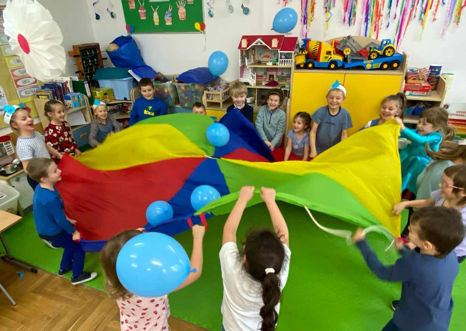  Zdjęcie przedstawia grupę dzieci, które bawią się wspólnie przy użyciu balonów i chusty animacyjnej.