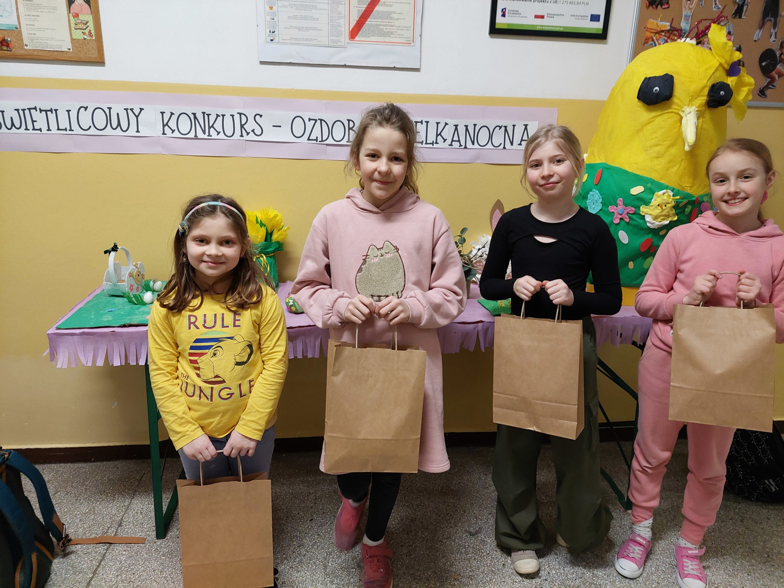 Cztery dziewczynki stoją na szkolnym korytarzu, w rękach trzymają papierowe torby z prezentami. Za nimi widać stół z ozdobami świątecznymi i napis na ścianie: świetlicowy konkurs - ozdoba wielkanocna. 