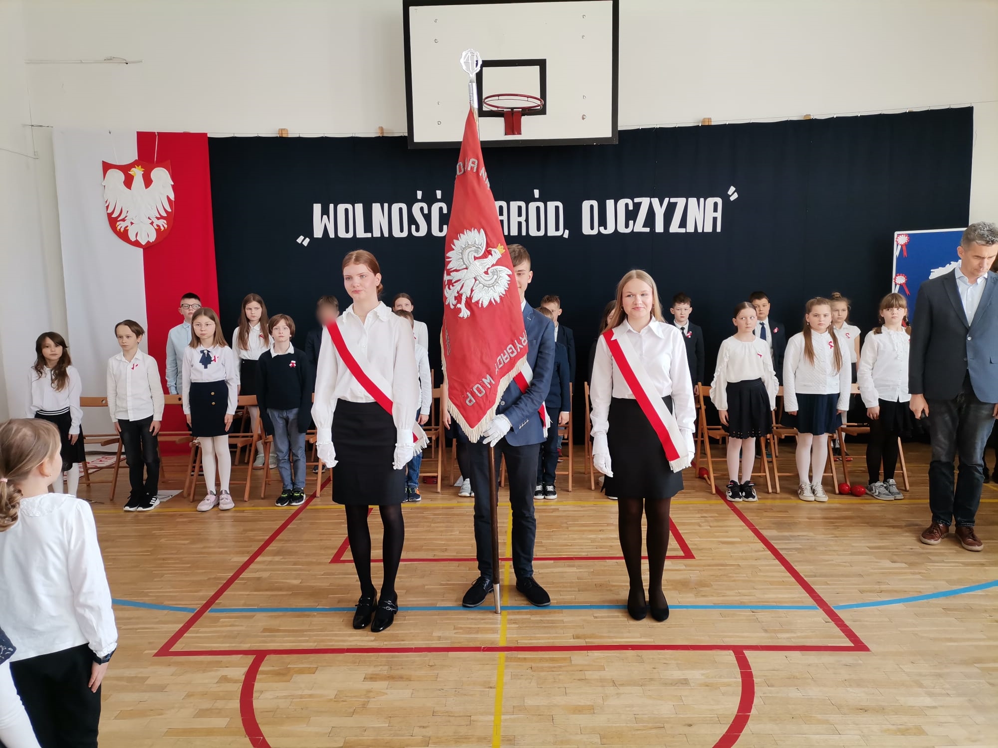 Duża grupa dzieci ubranych na galowo. Na pierwszym planie 2 dziewczyny i chłopiec pośrodku, który trzyma sztandar szkoły. W tle dekoracja, po jej lewej stronie flaga Polski i godło, na środku napis Wolność naród ojczyzna
