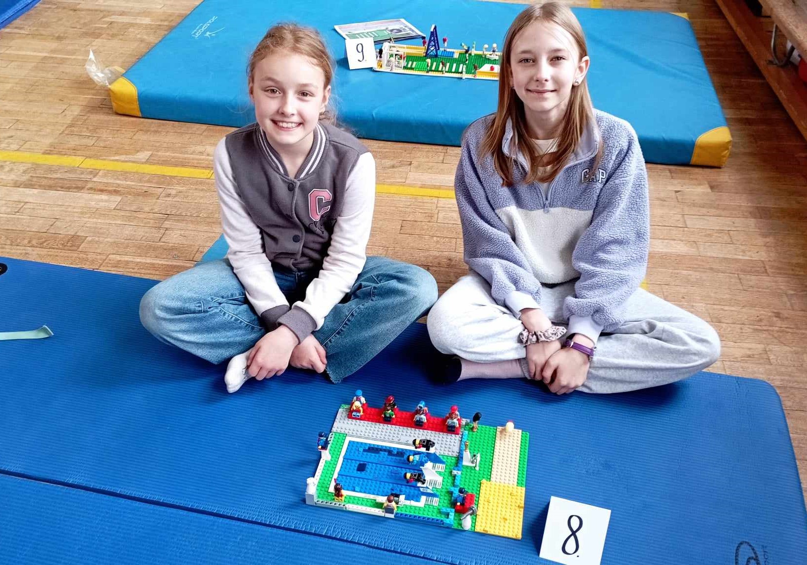 W sali gimnastycznej na niebieskiej karimacie siedzą dwie uczennice z klasy III. Przed nimi stoi ich budowla z klocków LEGO i etykietka z cyfrą 8. W tle inny materac z budowlą z klocków.
