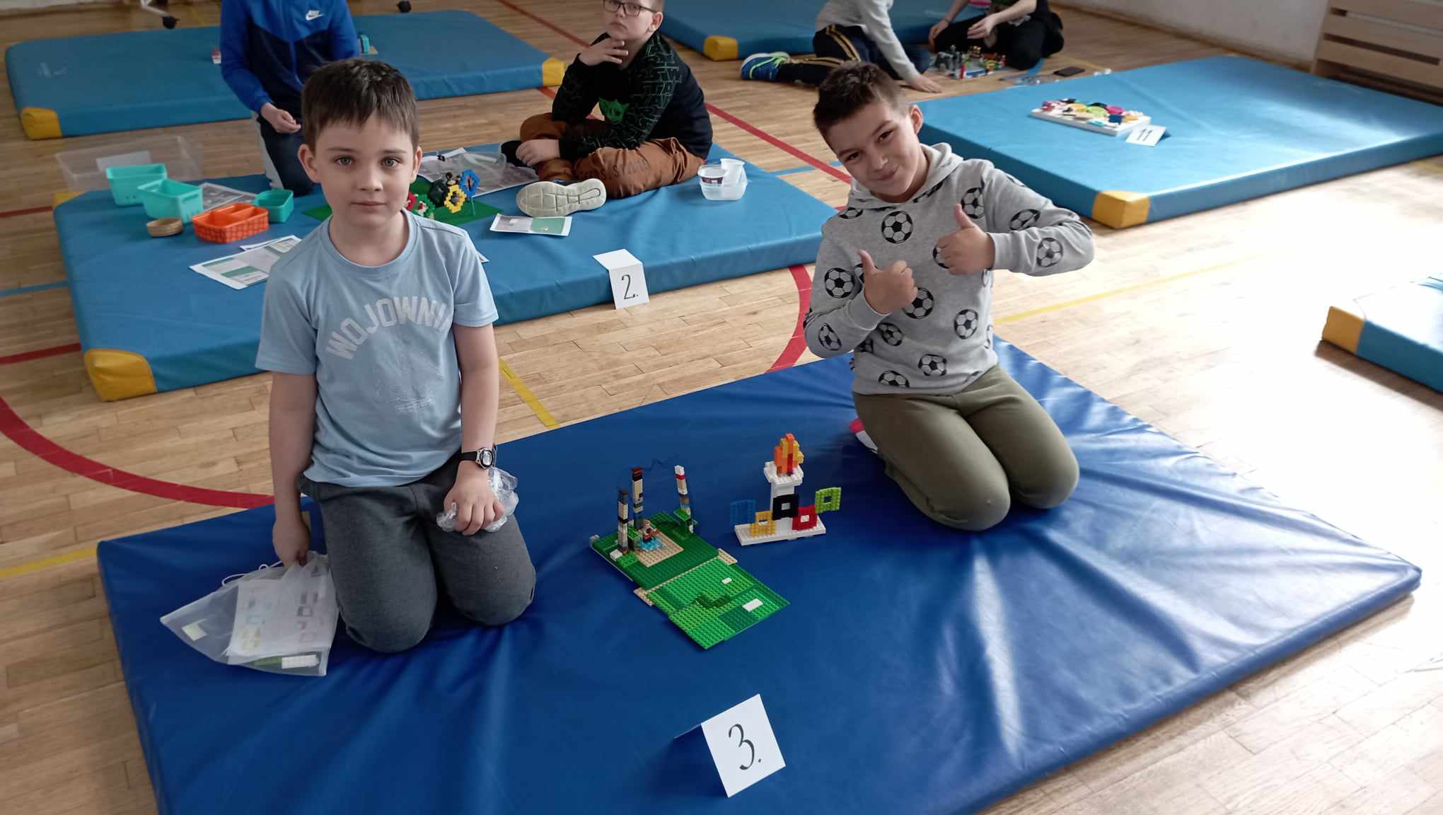 W sali gimnastycznej na niebieskim materacu siedzi dwoje uczniów z klasy II. Przed nimi stoi ich budowla z klocków LEGO i etykietka z cyfrą 3. W tle 5 innych materacy z dziećmi i budowlami z klocków.
