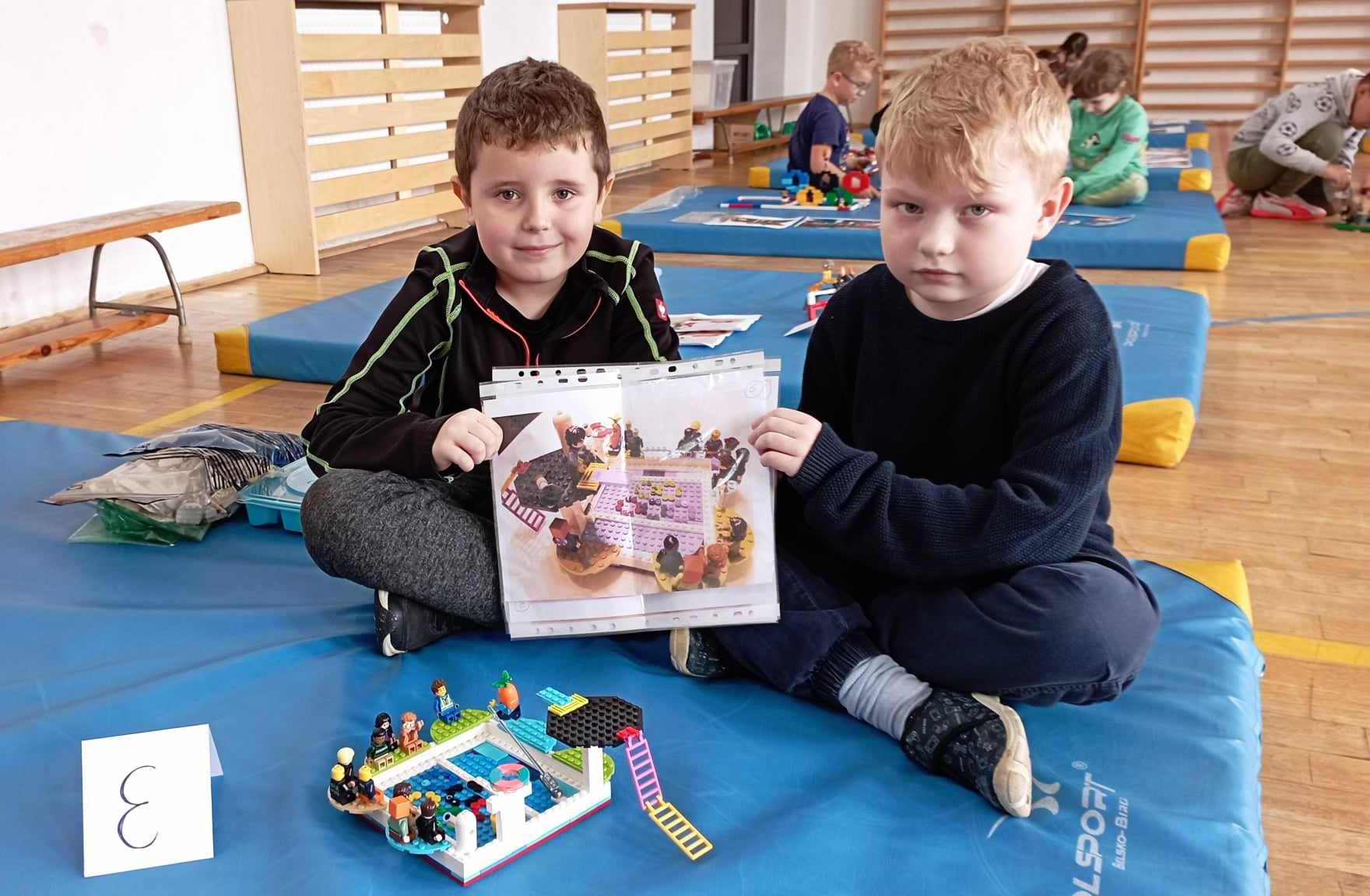 W sali gimnastycznej na niebieskim materacu siedzi dwoje chłopców z klasy 0. Przed nimi stoi ich budowla z klocków LEGO i etykietka z literą E. W tle 4 inne materace z dziećmi i budowlami z klocków.