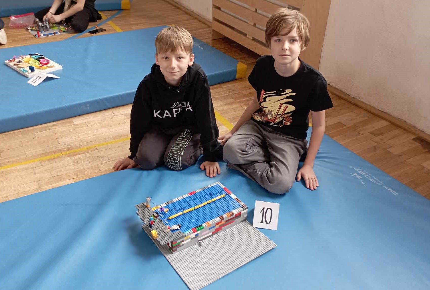 W sali gimnastycznej na niebieskim materacu siedzi dwoje uczniów z klasy III. Przed nimi stoi ich budowla z klocków LEGO i etykietka z liczbą 10. W tle inny materac z dziećmi i budowlami z klocków.