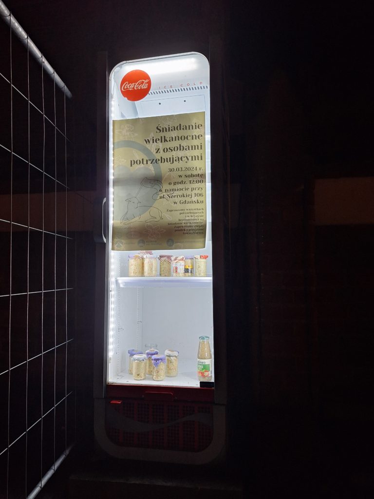Zdjęcie przedstawia wielką lodówkę, na której umieszczony jest plakat informujący o śniadaniu wielkanocnym z osobami potrzebującymi. W lodówce stoją słoiki z sałatką.