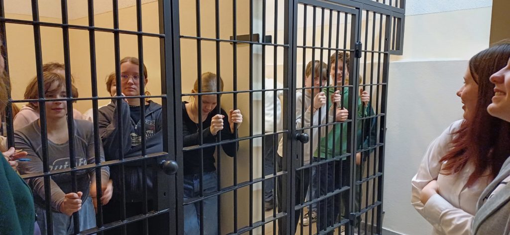 Uczniowie stoją zamknięci za kratami w areszcie. W jednym z nich znajdują dziewczynki, a w drugim chłopcy.