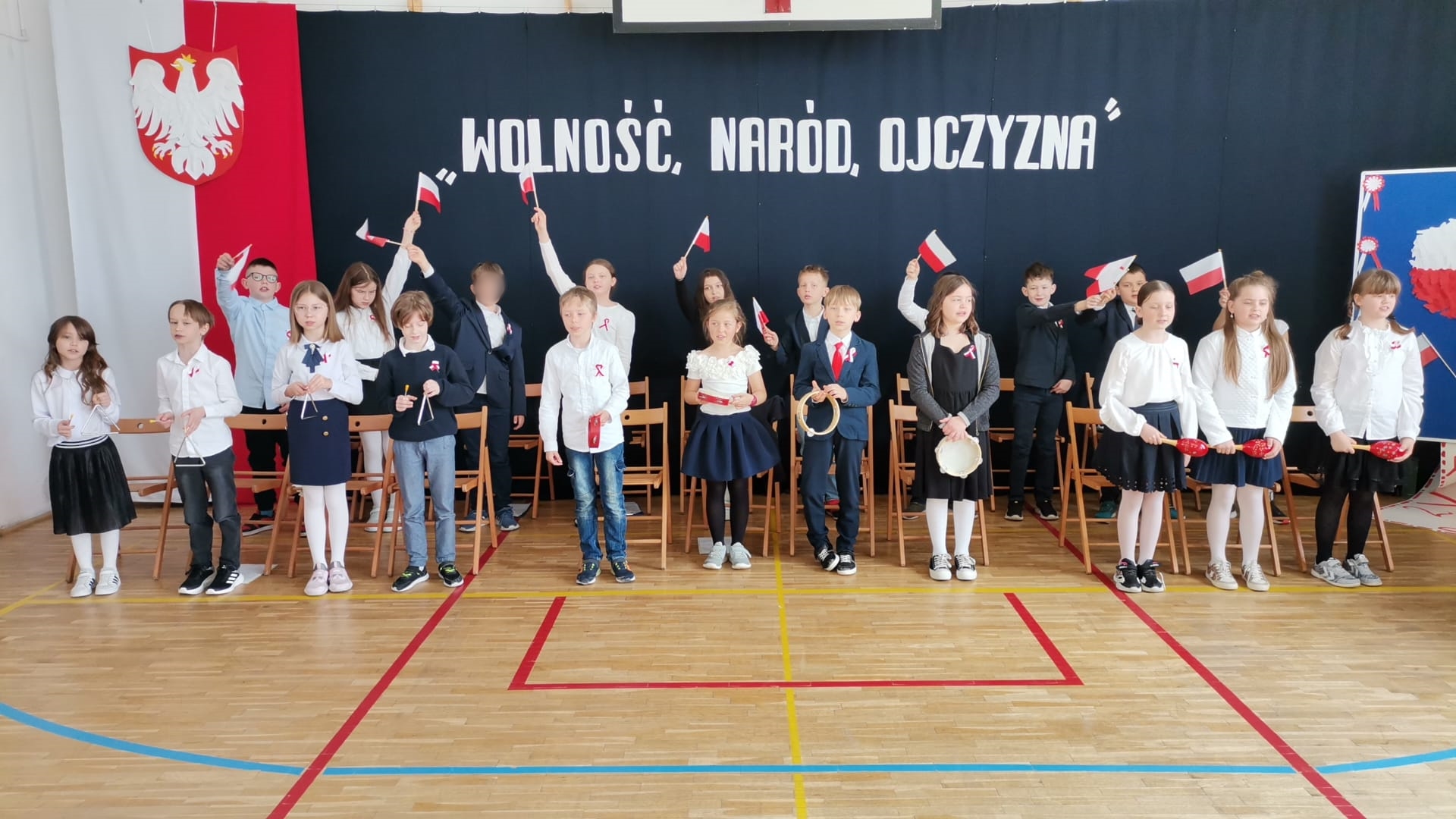 22 uczniów w dwóch rzędach. Z przodu dzieci trzymają instrumenty, rząd drugi z tyłu, dzieci machają flagami Polski. W tle dekoracja, po jej lewej stronie flaga Polski i godło, na środku napis Wolność naród ojczyna