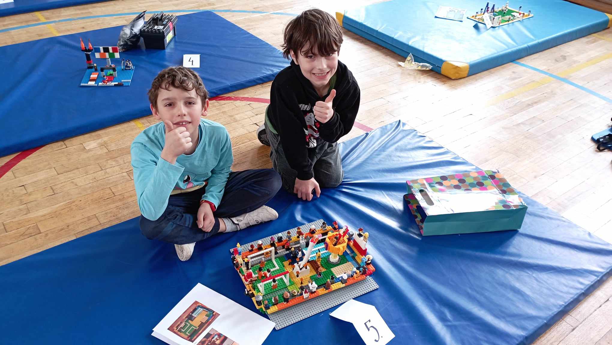 W sali gimnastycznej na niebieskim materacu siedzi dwoje uczniów z klasy II trzymających uniesiony w górę kciuk. Przed nimi stoi ich budowla z klocków LEGO i etykietka z cyfrą 5, obok leżą kartki z projektem budowli.