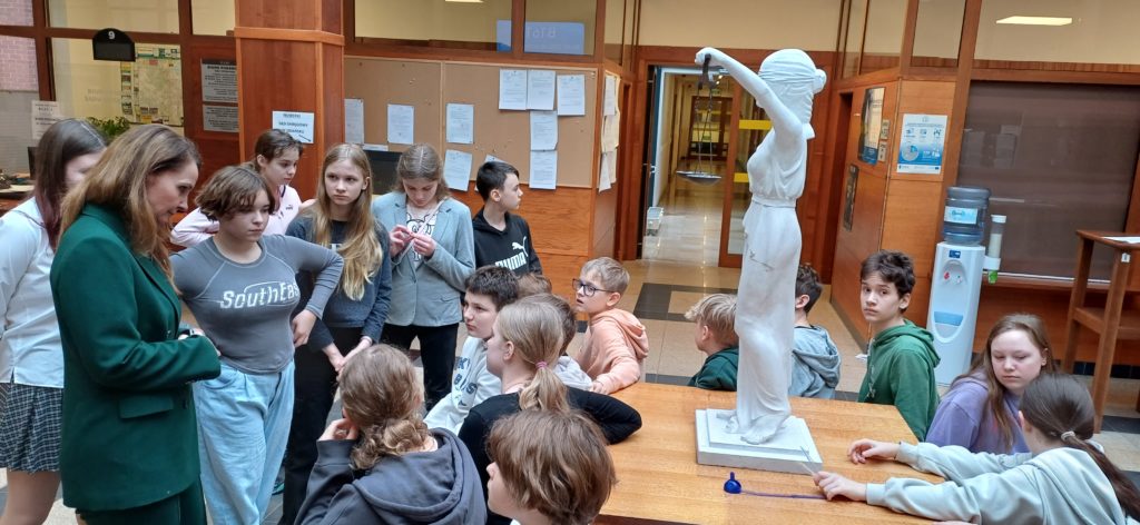 Część uczniów z klasy 6B siedzi, a pozostali stoją dookoła posągu greckiej bogini Temidy. Wśród nich stoi kobieta w zielonym ubraniu.