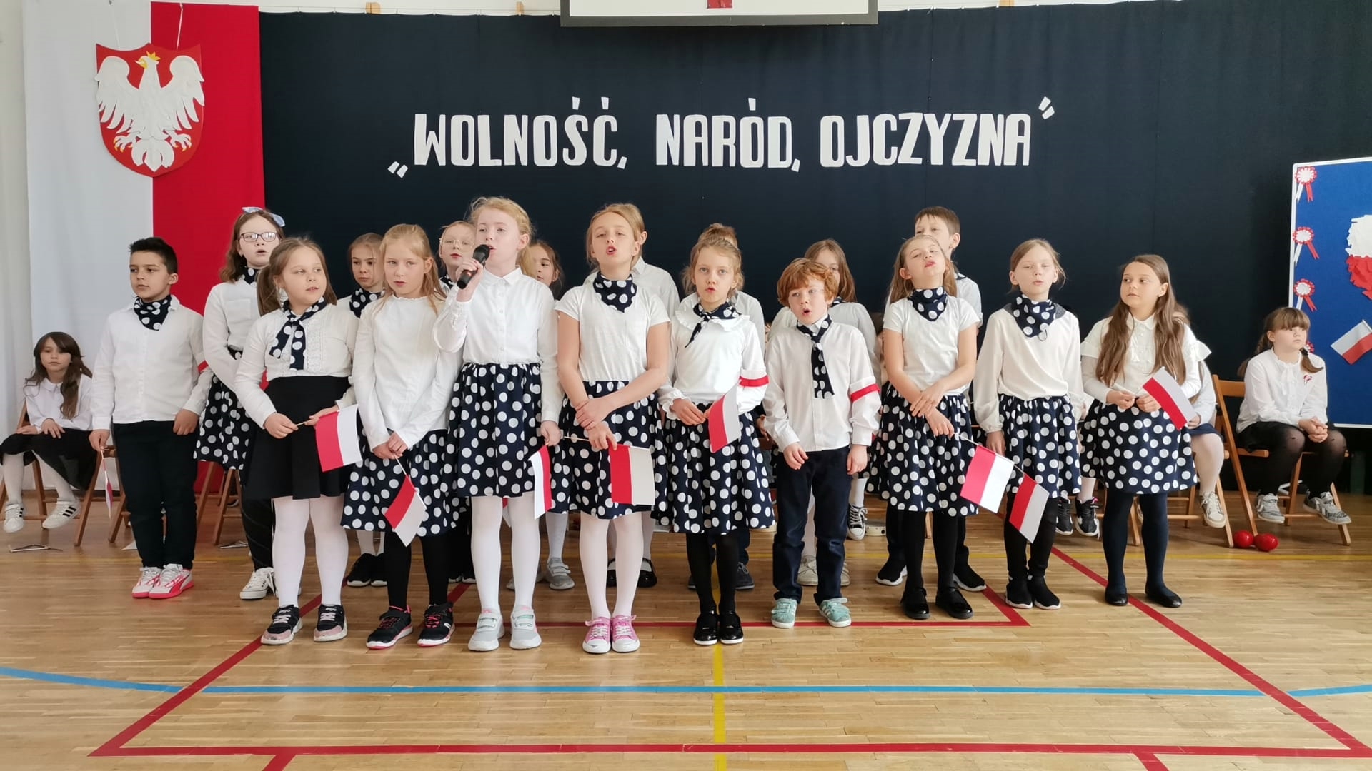  Grupa dzieci ubranych w jednakowe stroje, dziewczynki w spódnice z groszkami, wszyscy z białymi koszulami. Jedna z dziewcząt trzyma mikrofon. W tle dekoracja, po jej lewej stronie flaga Polski i godło, na środku napis Wolność naród ojczyna.