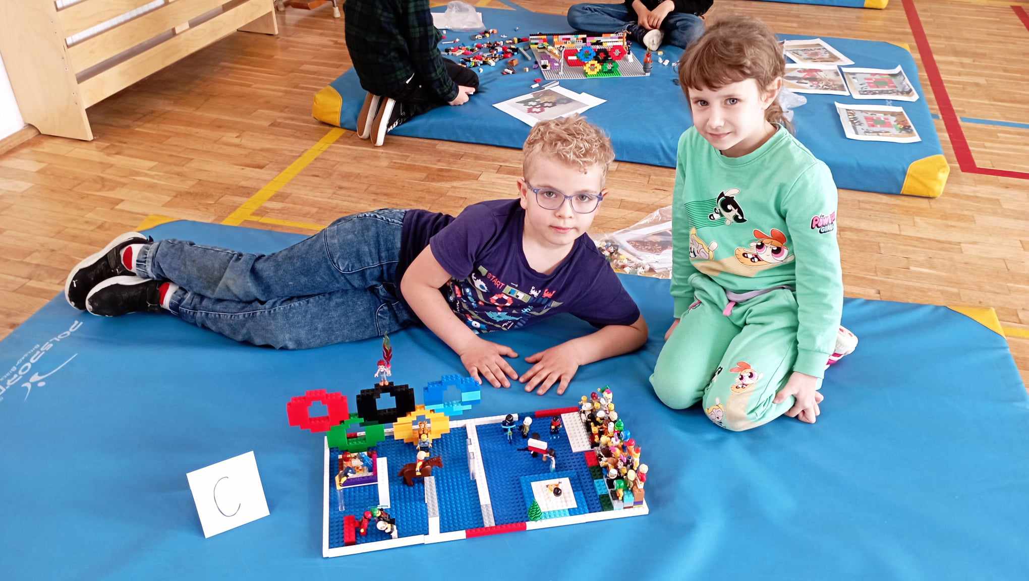 W sali gimnastycznej na niebieskim materacu leży chłopczyk, obok siedzi dziewczynka z klasy 0. Przed nimi stoi ich budowla z klocków LEGO i etykietka z literą C. W tle inny niebieski materac z klockami LEGO i dwojgiem uczniów.