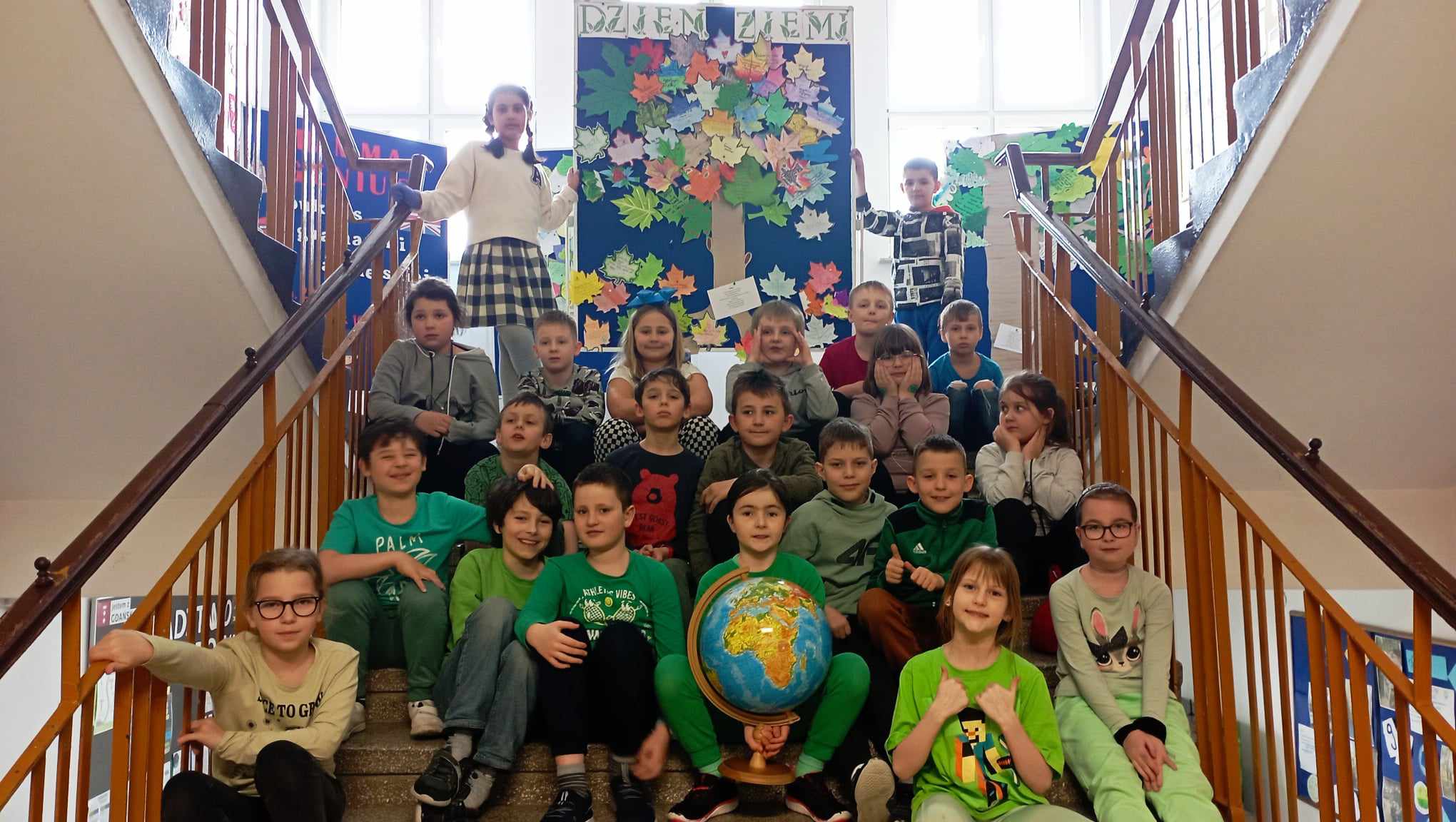 Grupa uśmiechniętych dzieci siedzi w trzech rzędach na schodach. Dziewczynka siedząca pośrodku pierwszego rzędu trzyma duży globus. W tle dwoje stojących dzieci trzyma tablicę przedstawiającą zrobione z papieru drzewo z liśćmi i napis "Dzień Ziemi".